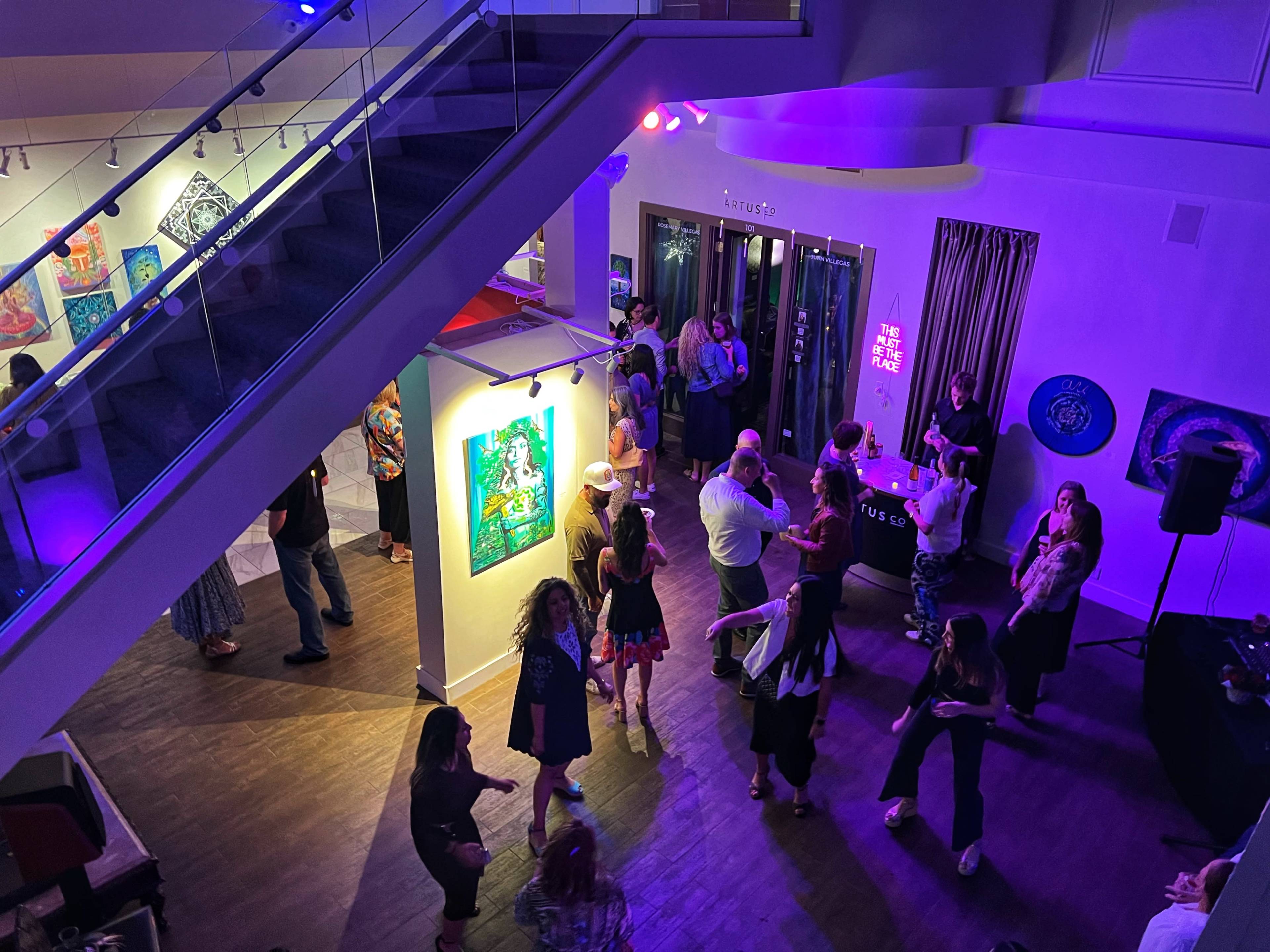 A vibrant gathering takes place in a modern art gallery, featuring people mingling, dancing, and enjoying the exhibits under colorful lighting.