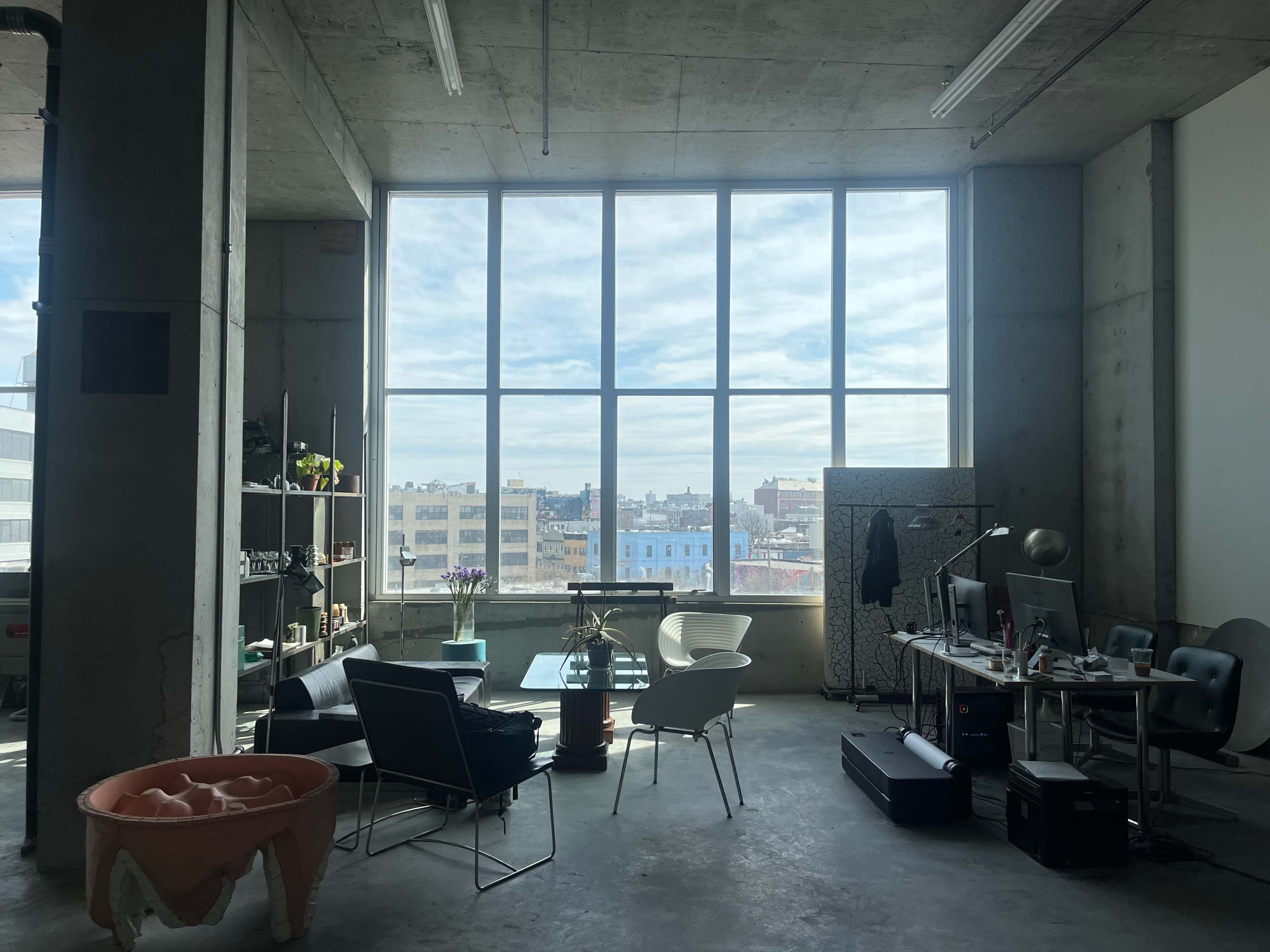 The image shows a modern office space with large windows, concrete walls, and a combination of furniture, including a glass table, chairs, and workstations.