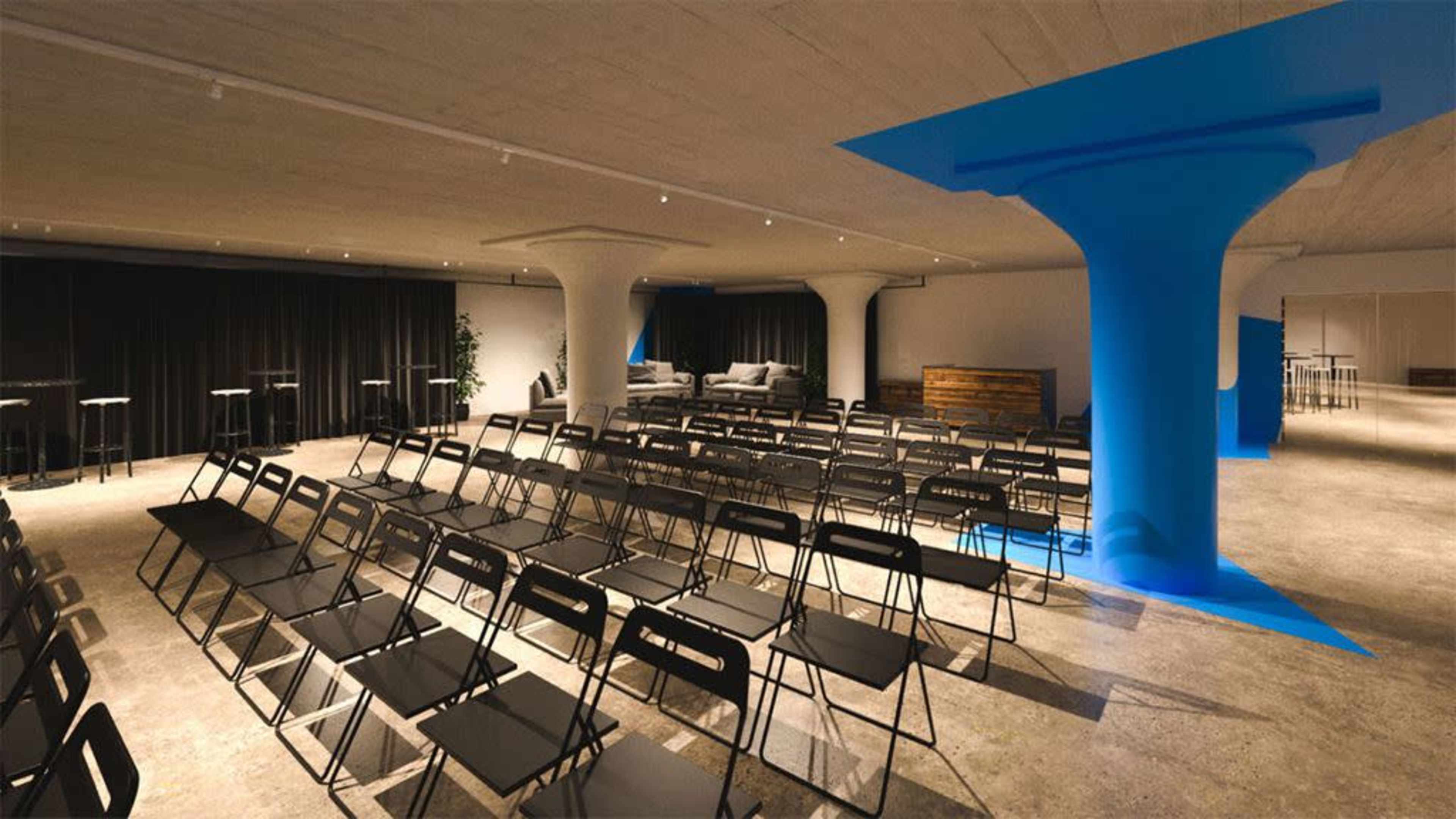 A modern event space with rows of black chairs facing a seating area, illuminated by natural light and featuring blue architectural highlights.