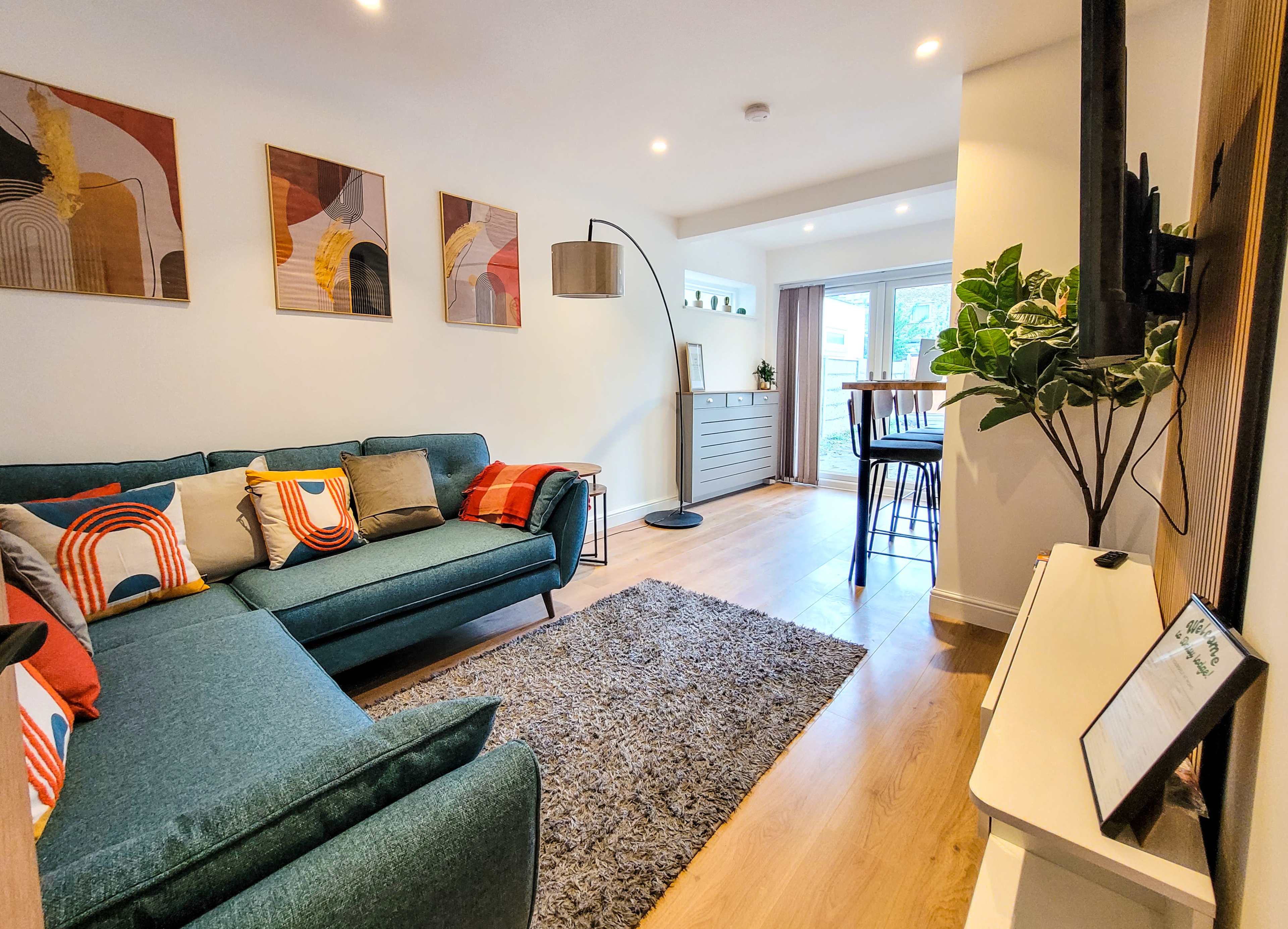 The scene shows a modern living room with a teal sofa, decorative pillows, a grey rug, wall art, and a dining area visible in the background.