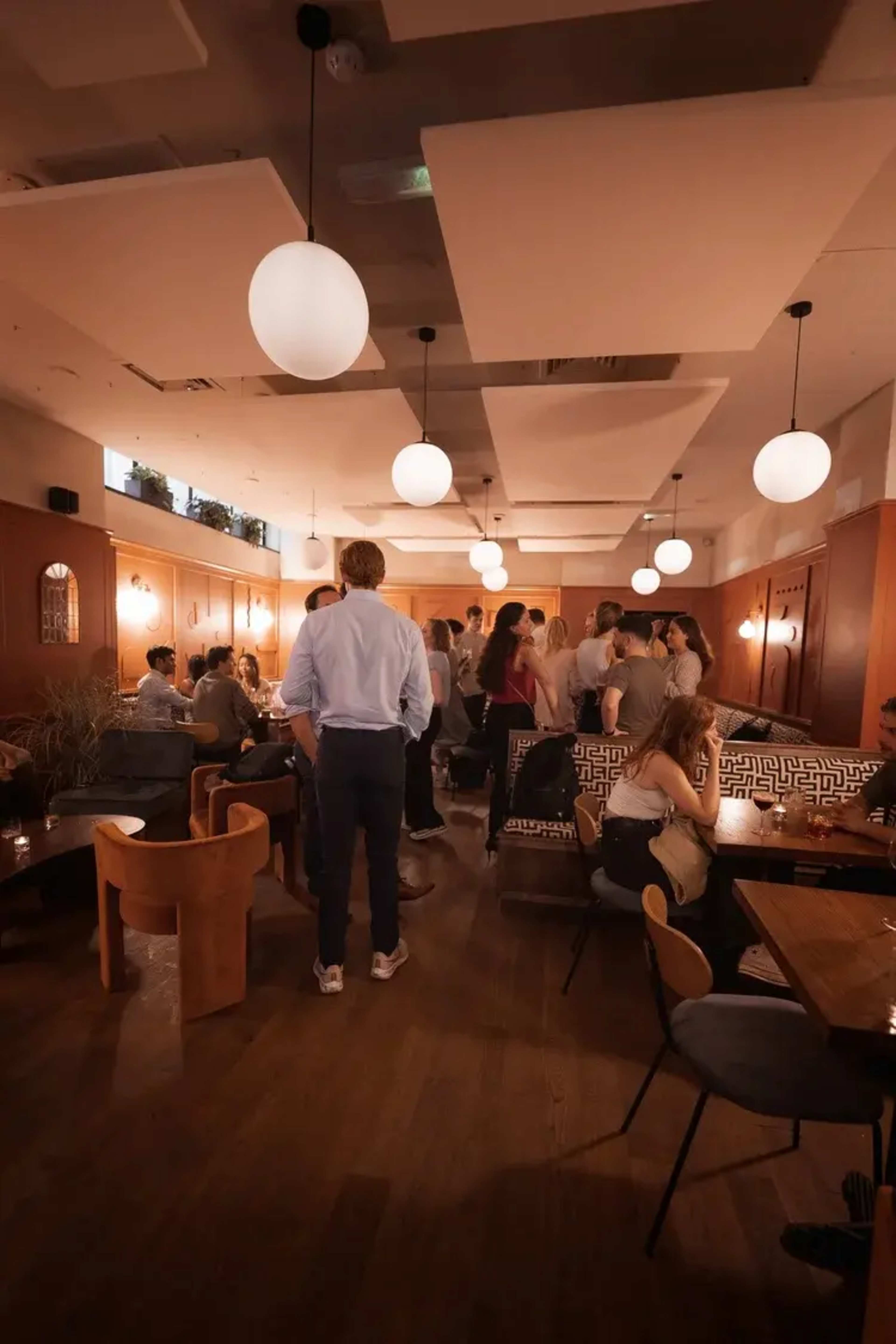 The image shows a bustling restaurant interior filled with patrons engaged in conversation, illuminated by round pendant lights.