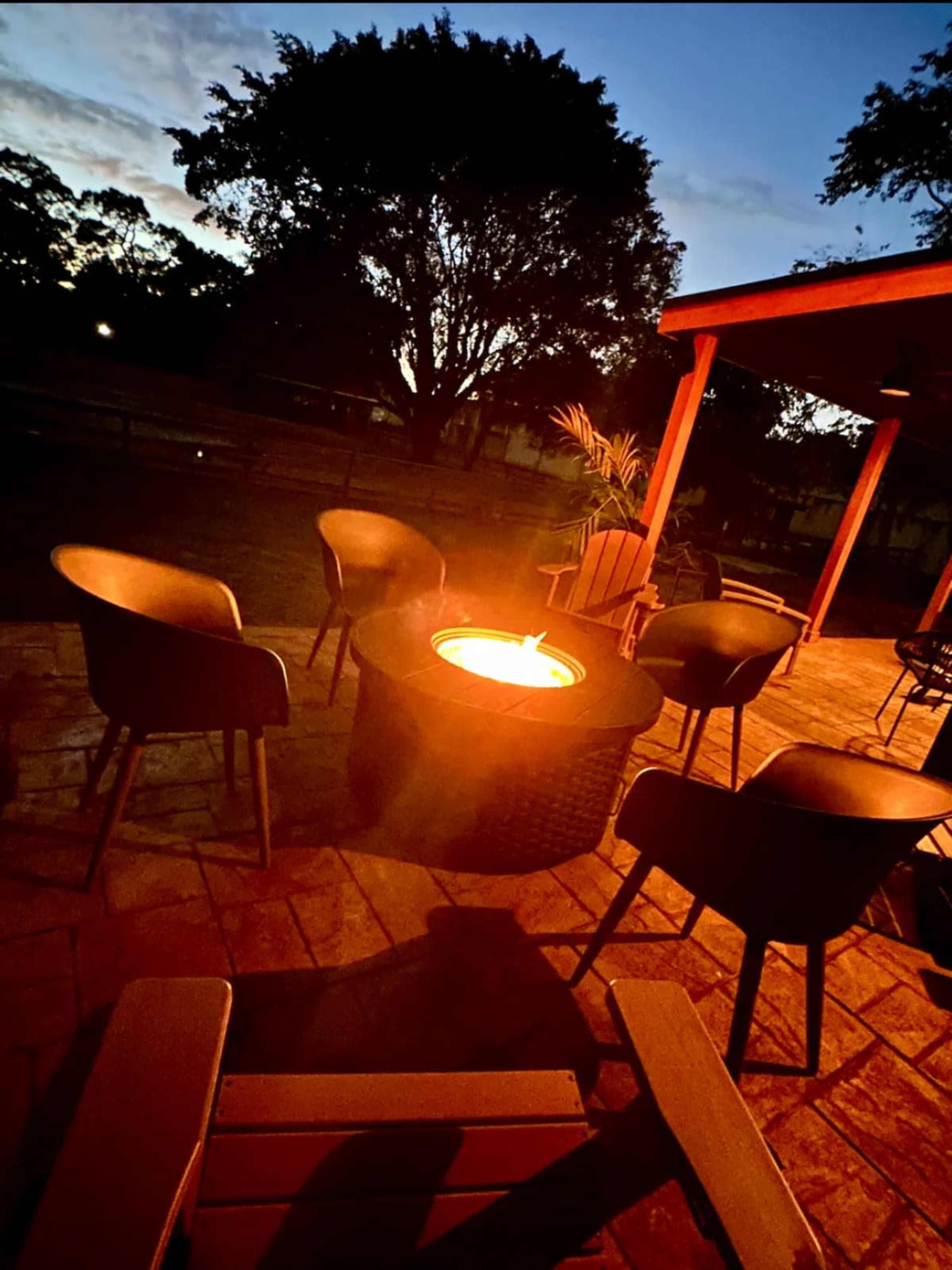 A fire pit surrounded by chairs is illuminated at dusk on a patio with trees in the background.