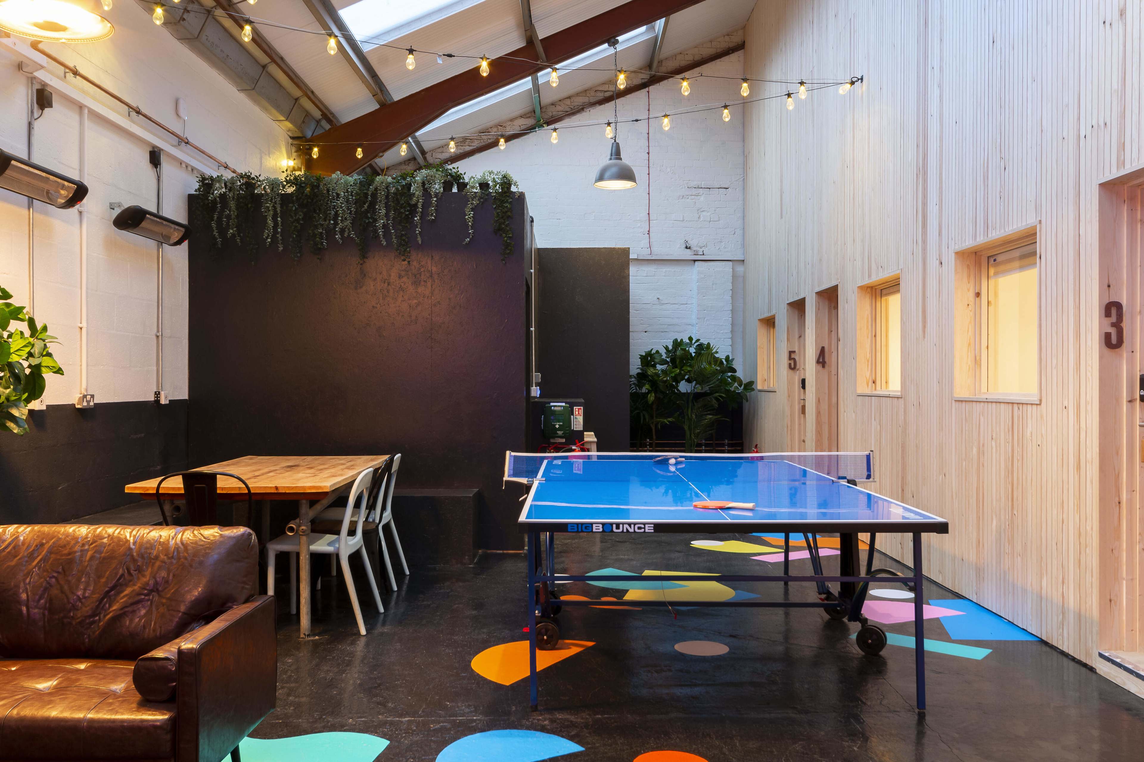 A brightly lit indoor space featuring a blue ping pong table, wooden seating, and several numbered rooms along the wall, decorated with plants and colorful floor designs.