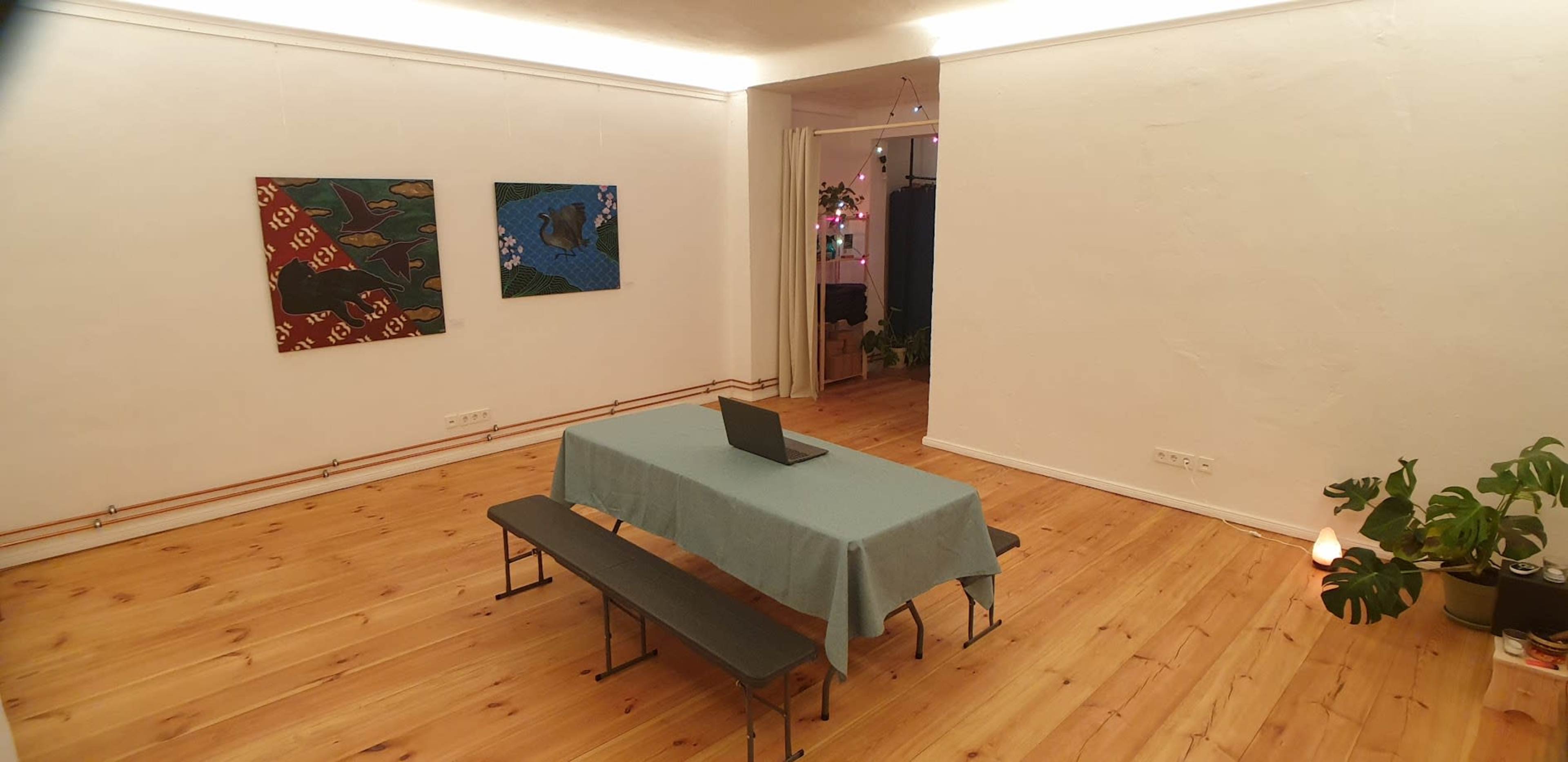 The image shows a minimalistic room with a table covered by a blue cloth, flanked by two benches, and adorned with two paintings on the wall and a potted plant in the corner.