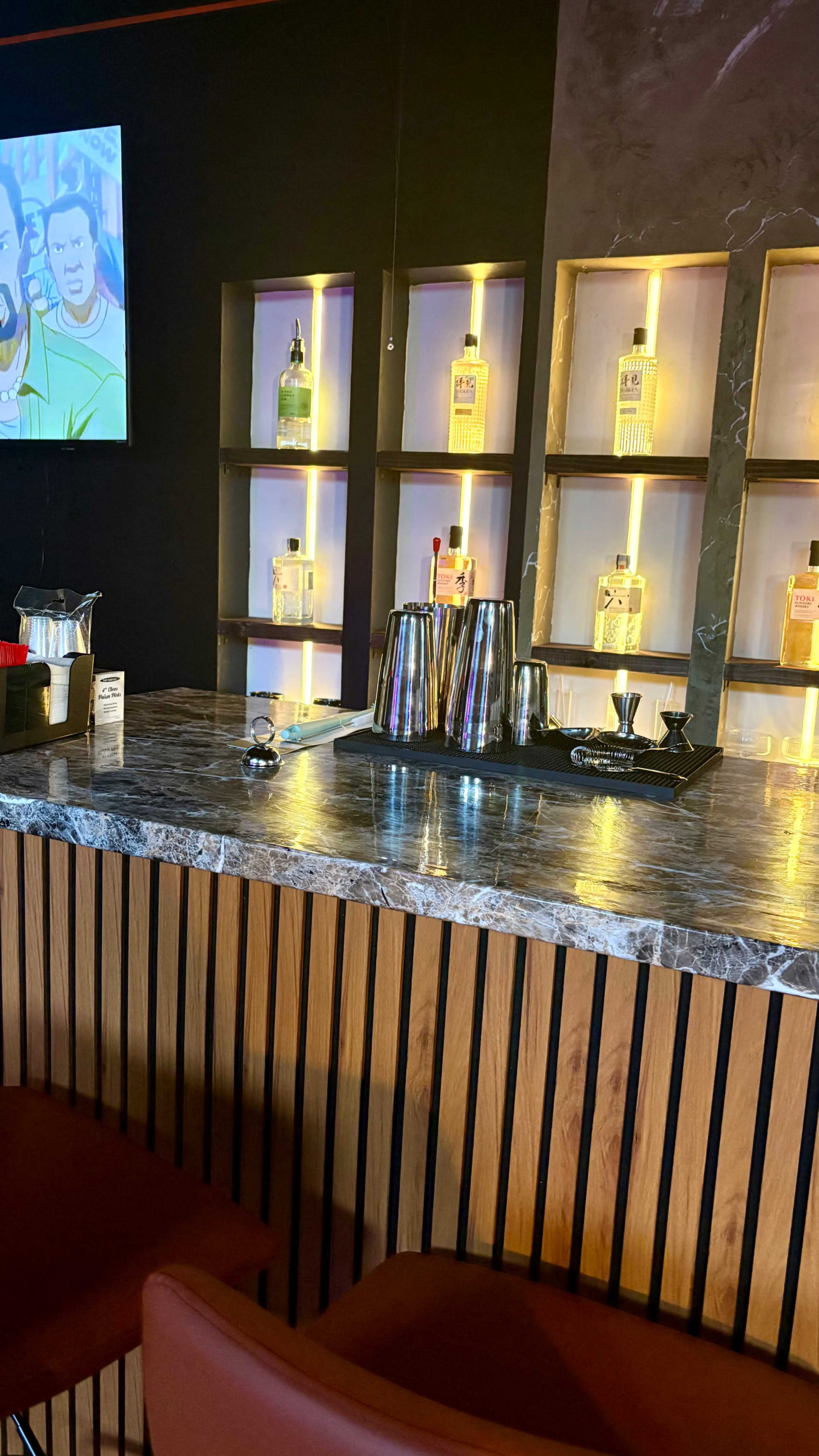 The image shows a bar with a marble countertop, featuring mixing tools and illuminated shelves displaying various bottles of liquor.