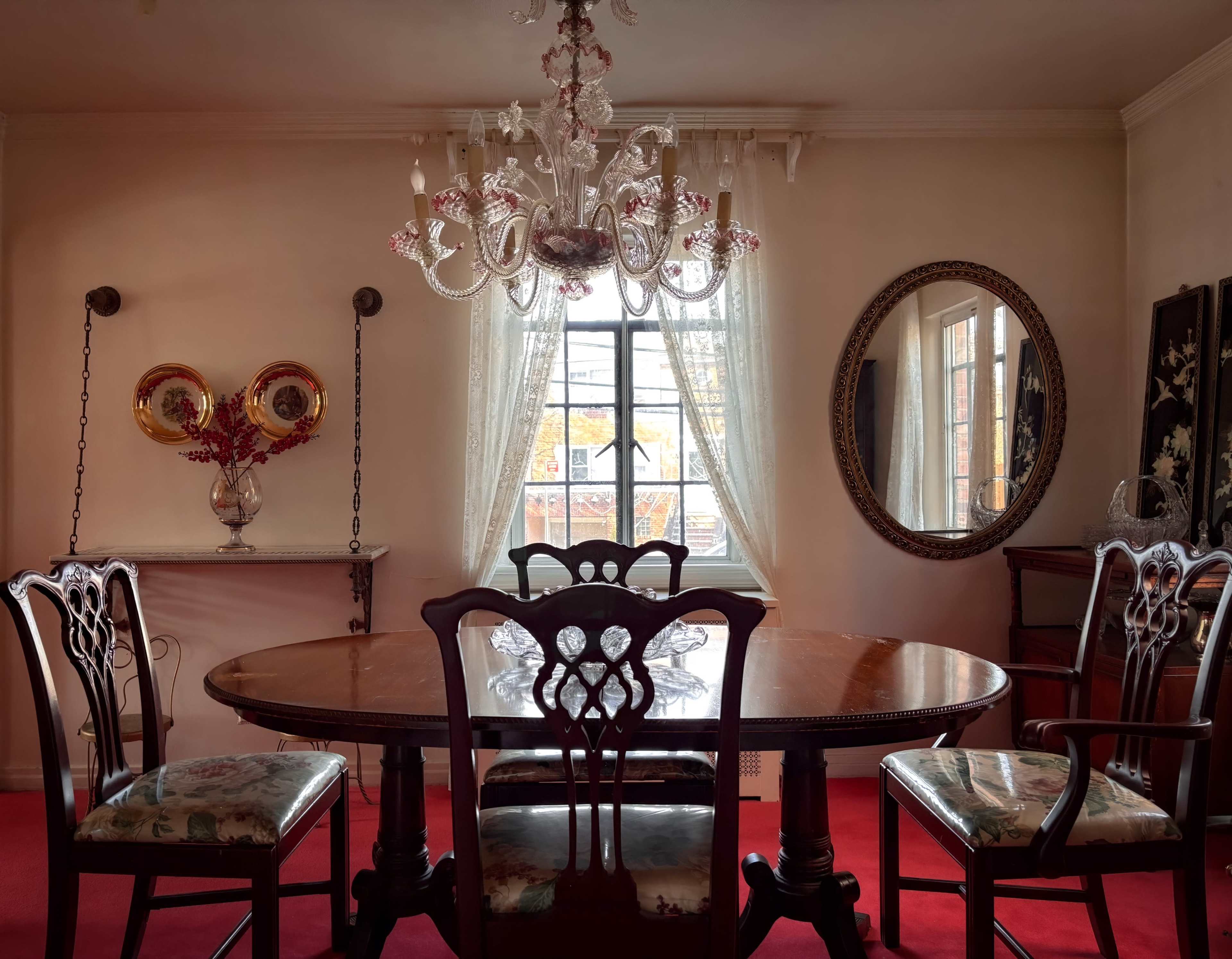 A formal dining room features a round wooden table surrounded by six chairs, with a chandelier overhead and a window providing natural light.
