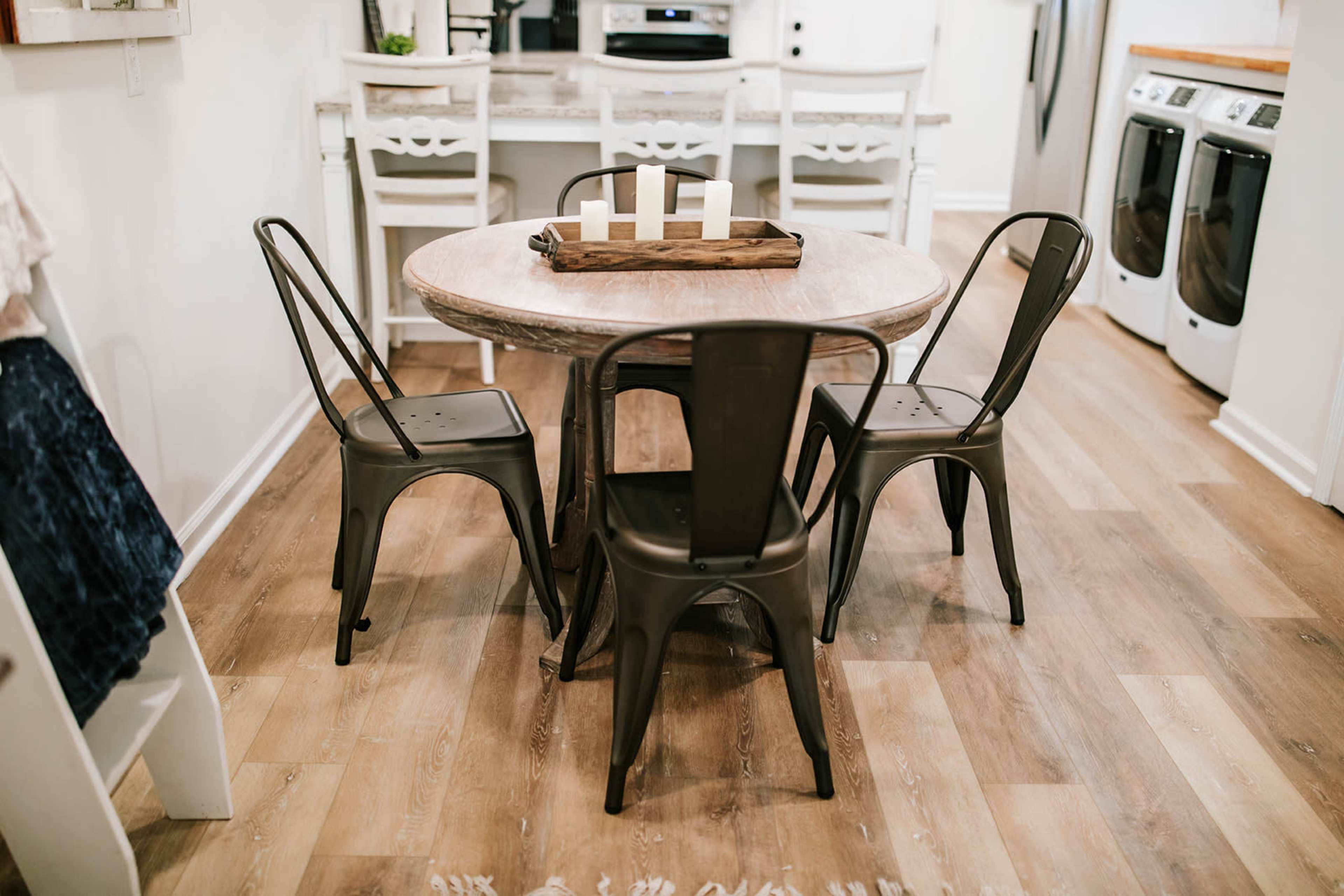 A round wooden dining table surrounded by four metal chairs is situated in a modern kitchen with light-colored flooring and appliances.