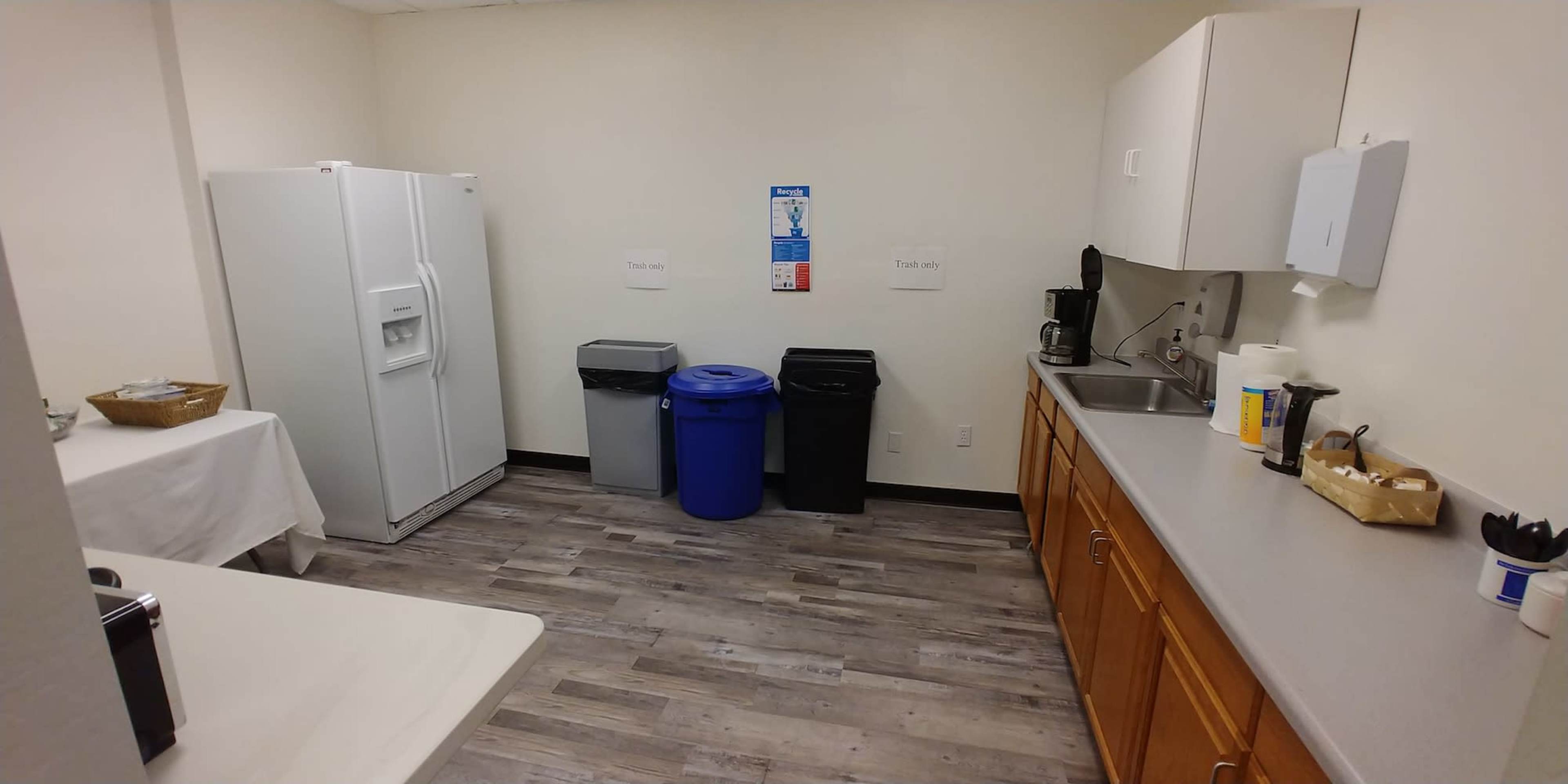 A break room with a refrigerator, a sink, a table with a cloth, and three trash bins arranged near the wall.