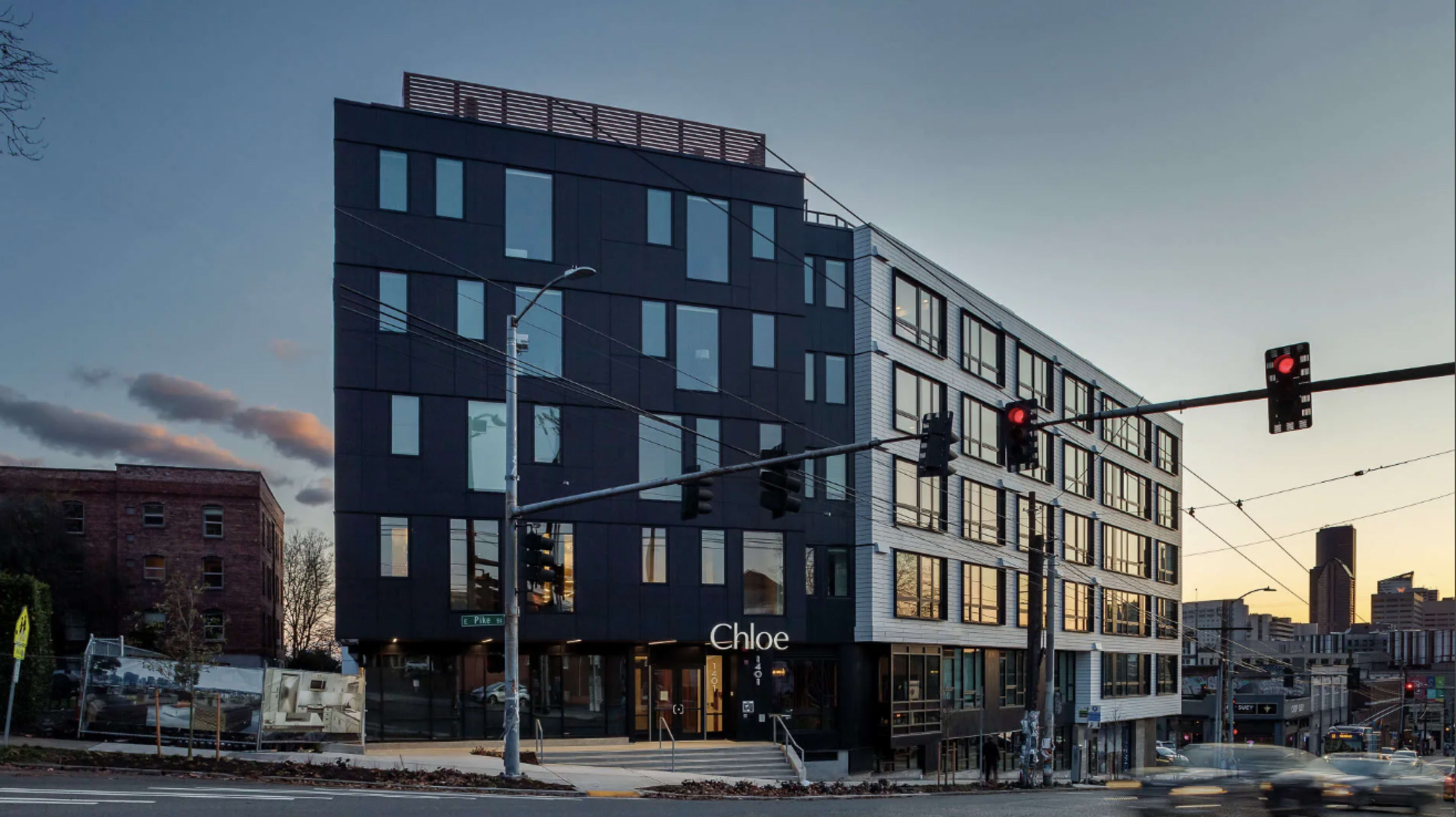 A modern building with a combination of black and white paneling, featuring large windows and a ground-level entrance marked "Chloe," sits at an intersection during twilight.