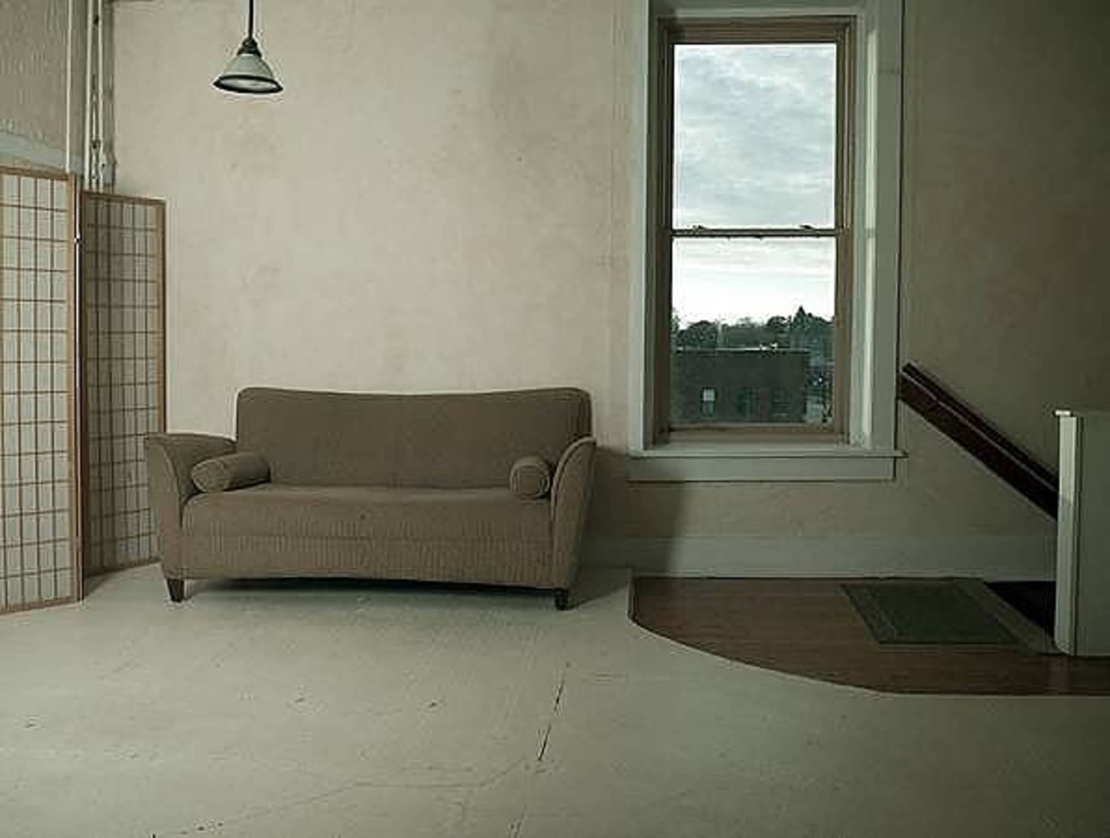 A sparsely furnished room with a brown couch, a window with cloudy skies outside, and a staircase in the background.