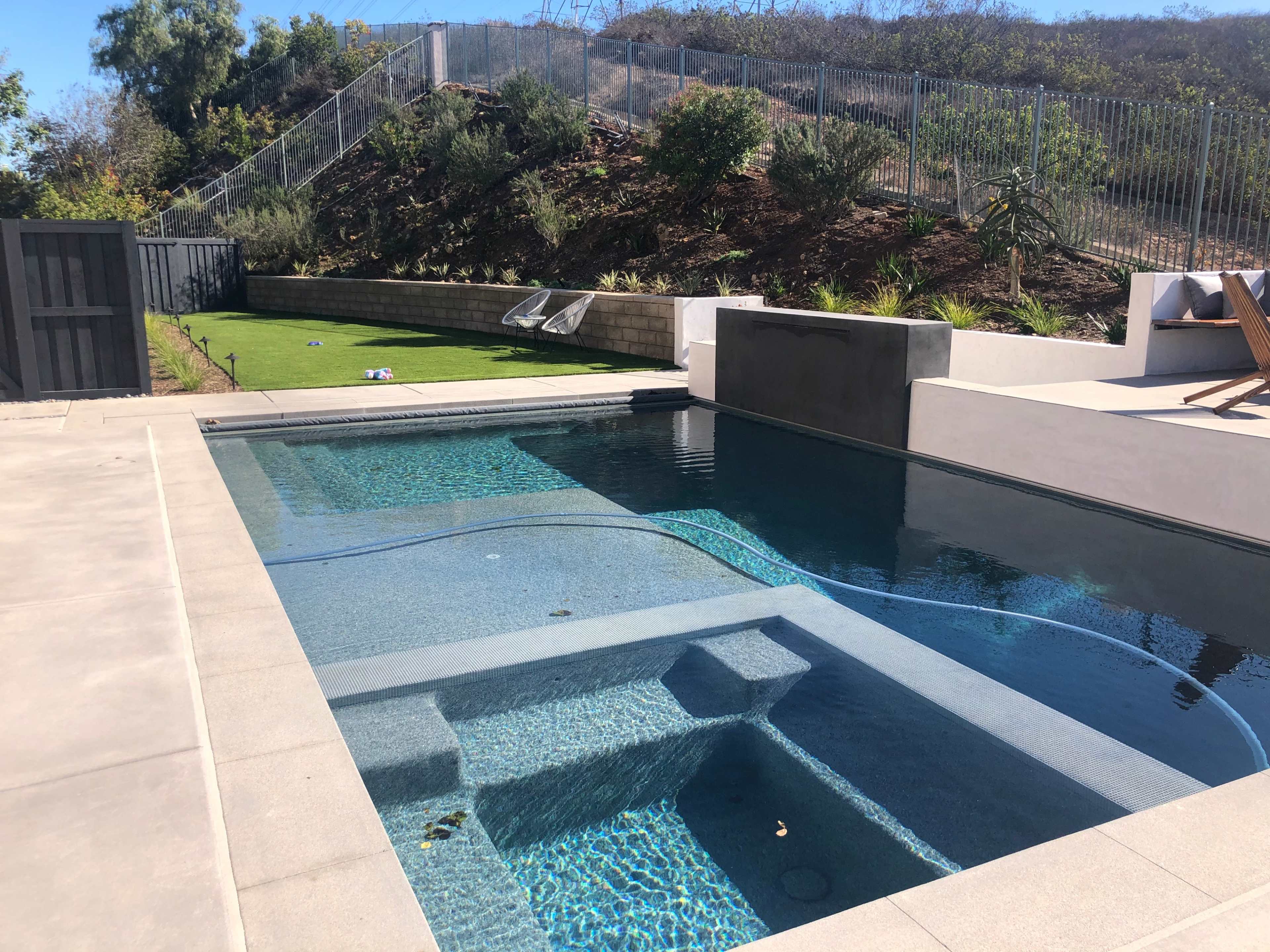 The image shows a swimming pool with a built-in spa, surrounded by a patio and landscaped hillside.