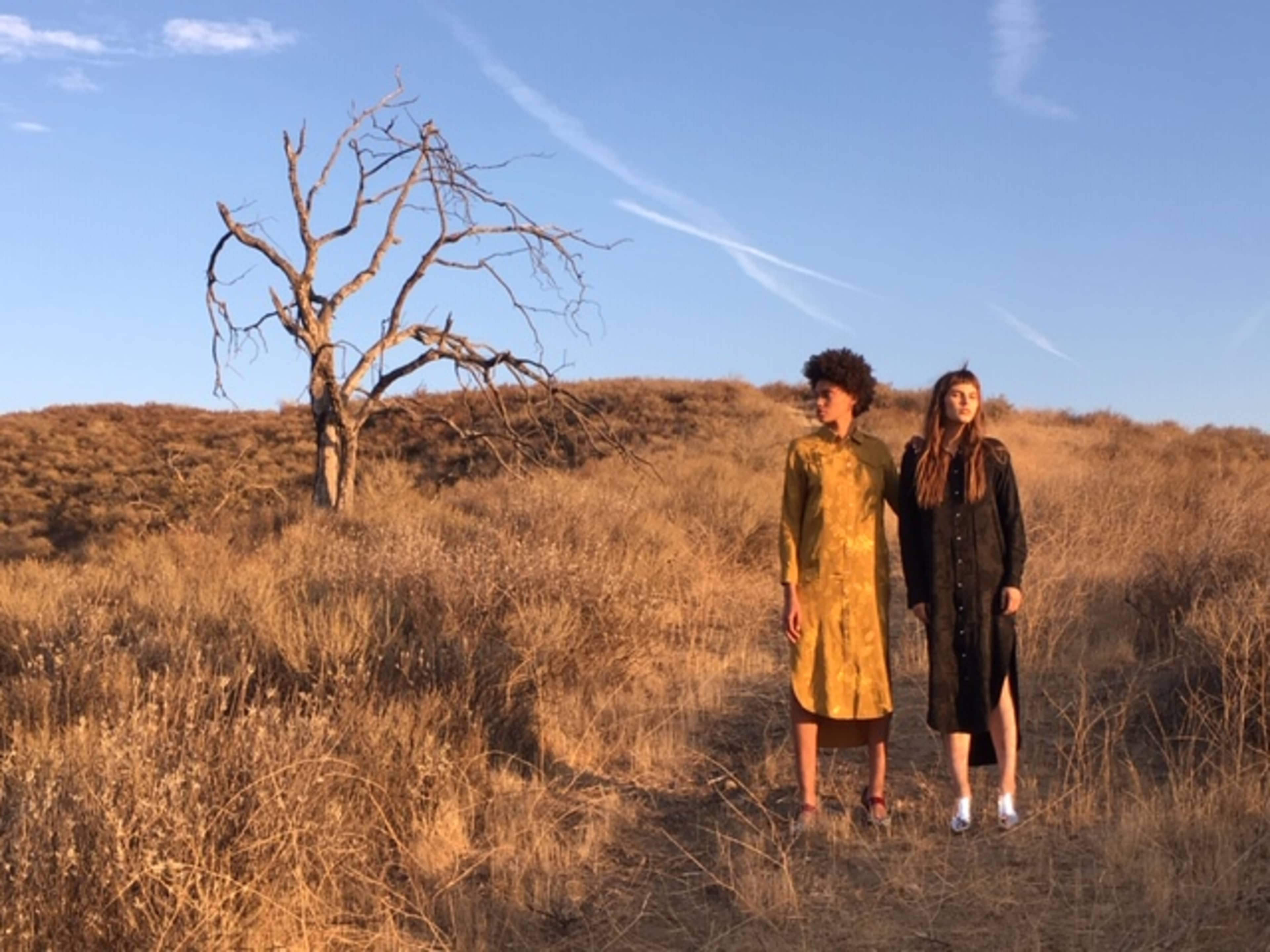 Two models stand on a dry, grassy hillside near a barren tree under a blue sky with wispy clouds.