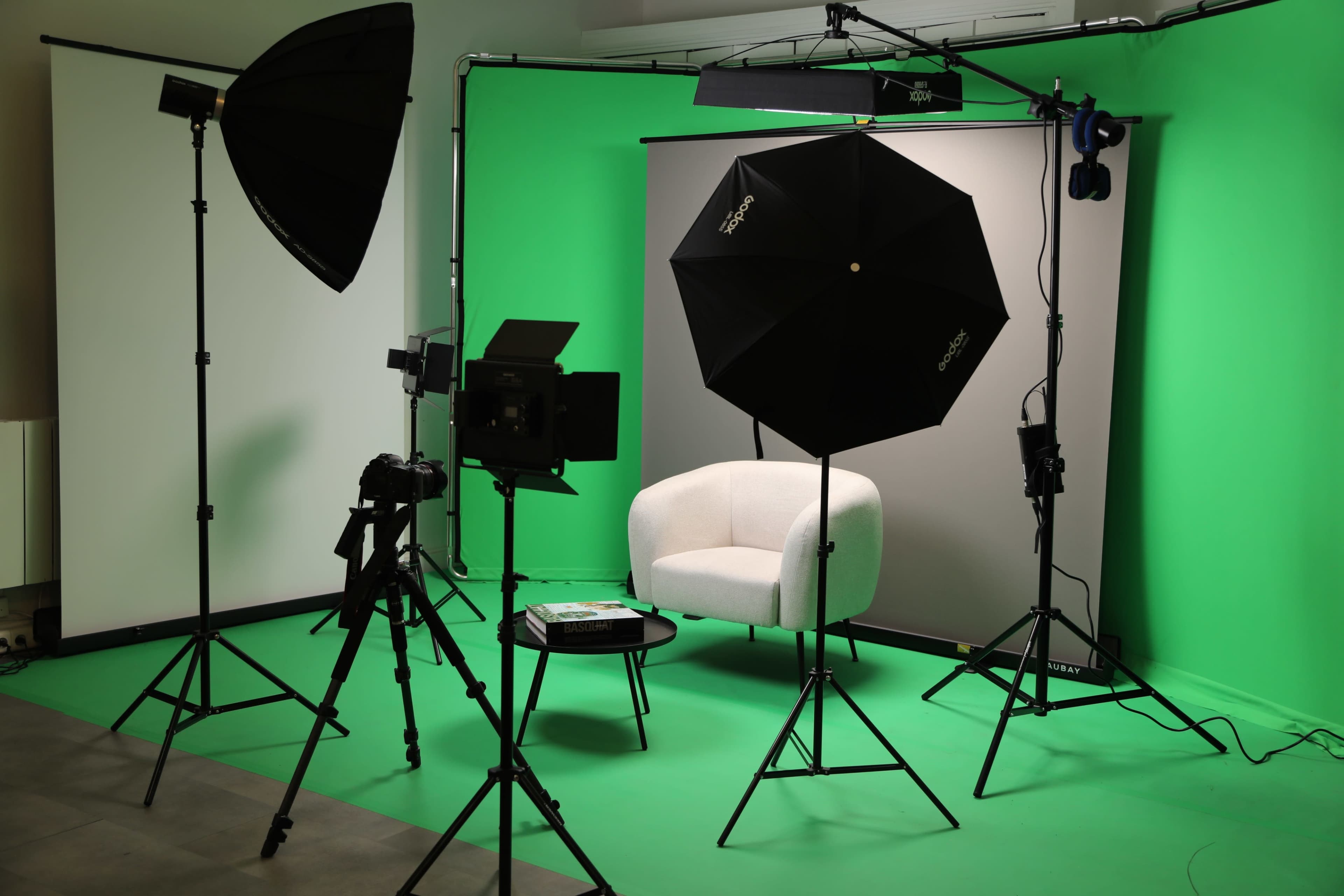 A studio setup featuring a white chair, backdrop, lighting equipment, and cameras on green flooring.