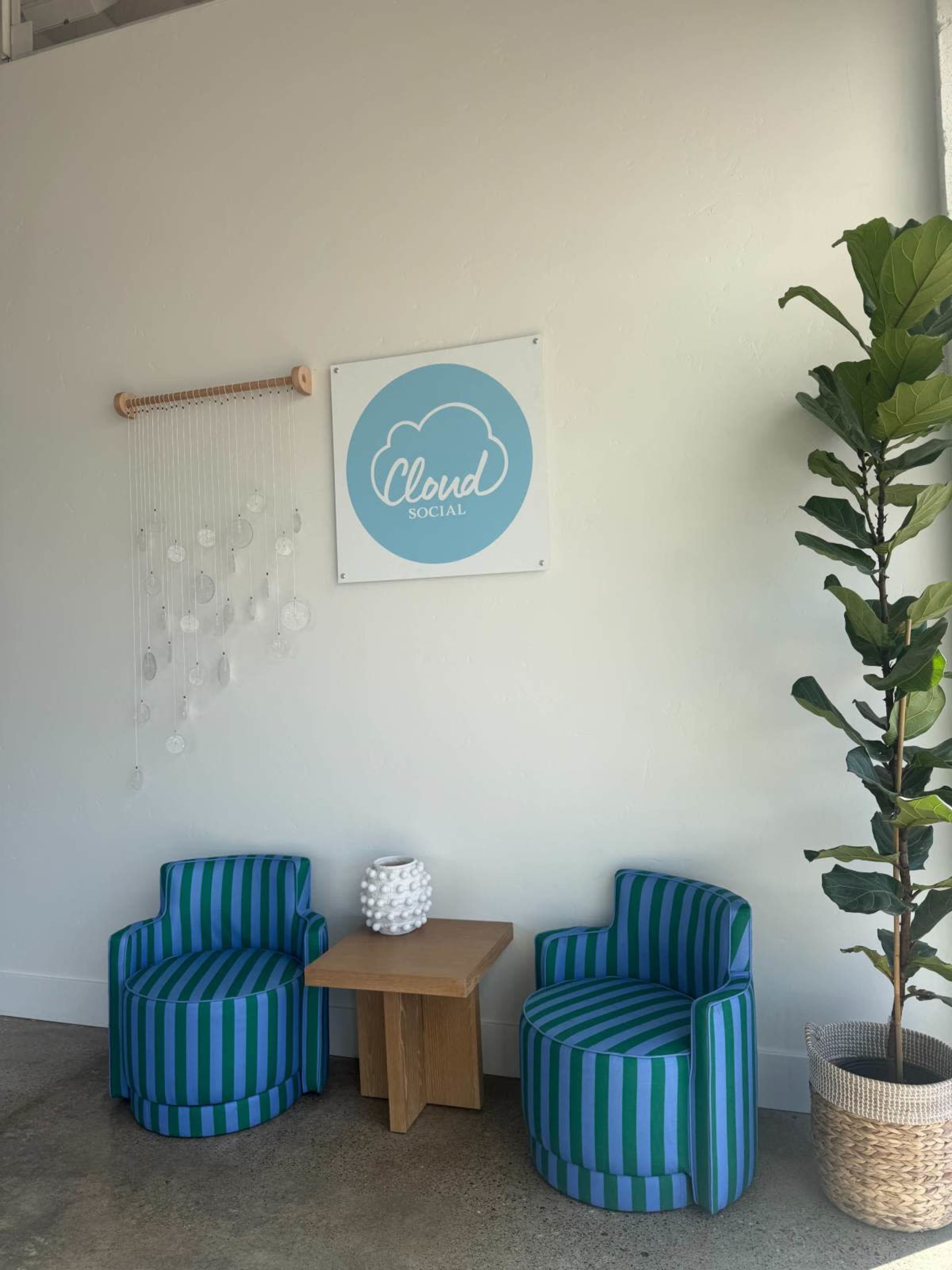 The image shows two striped blue and green chairs with a small wooden table in between them, against a white wall featuring a sign that reads "Cloud Social" and a decorative plant in a basket.
