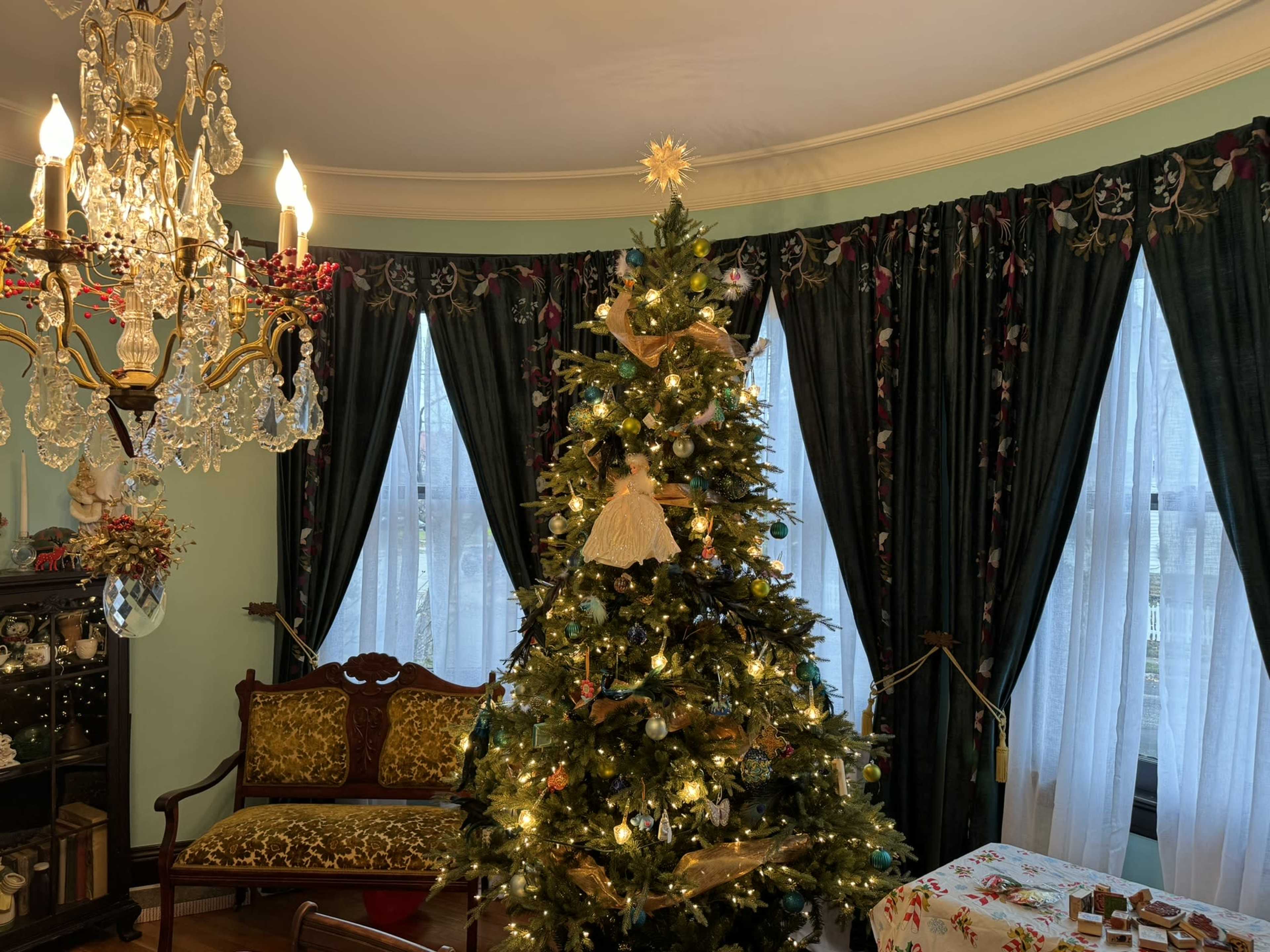 A decorated Christmas tree stands in a room with a chandelier, surrounded by curtains and a vintage sofa.