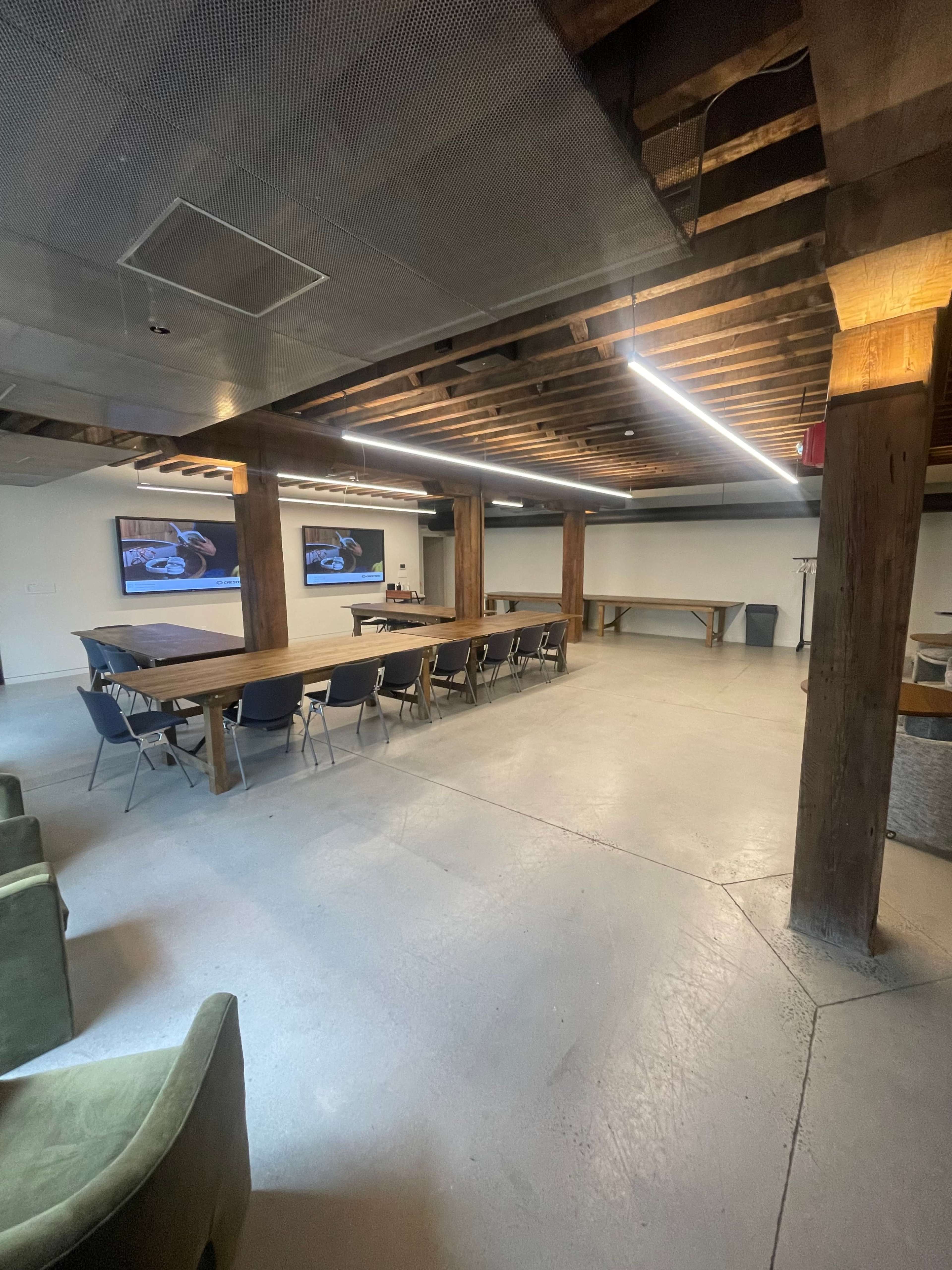 A spacious meeting room with wooden beams, a large table, rows of chairs, and two screens mounted on the walls.