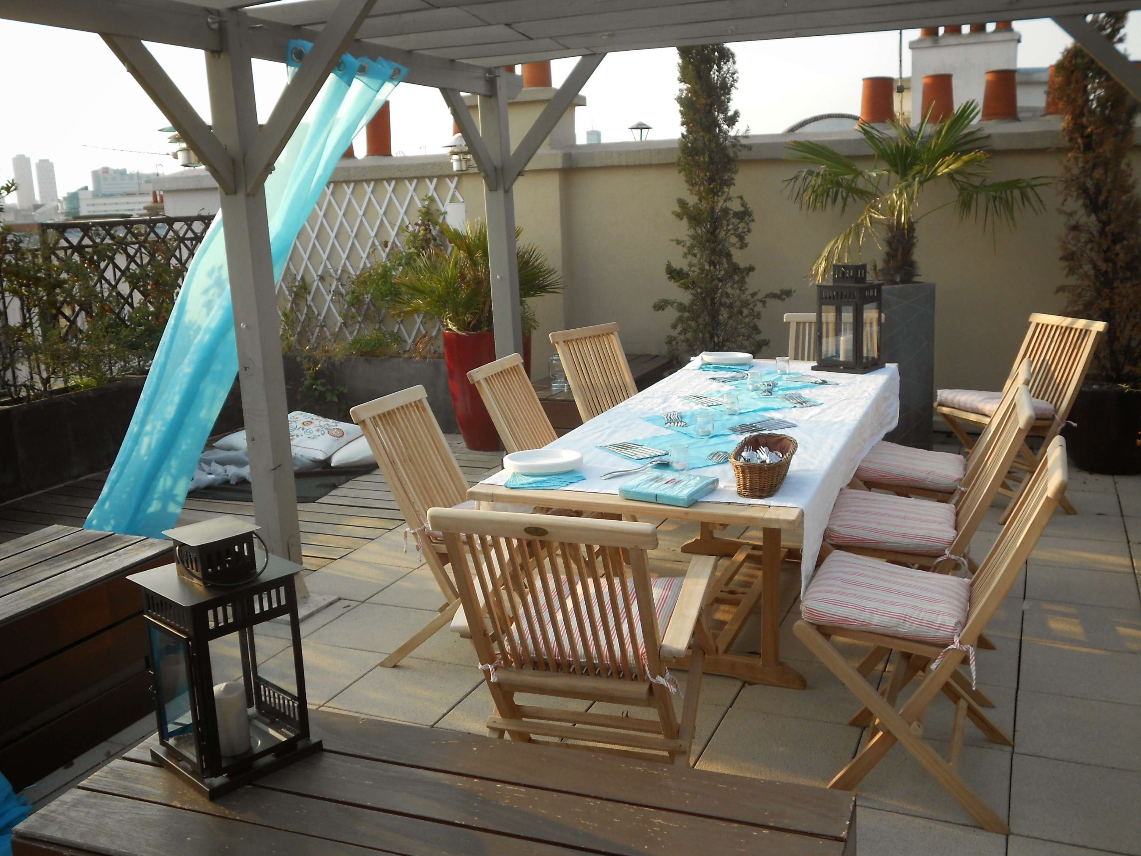 A wooden outdoor dining table is set with turquoise tableware under a pergola on a rooftop terrace.
