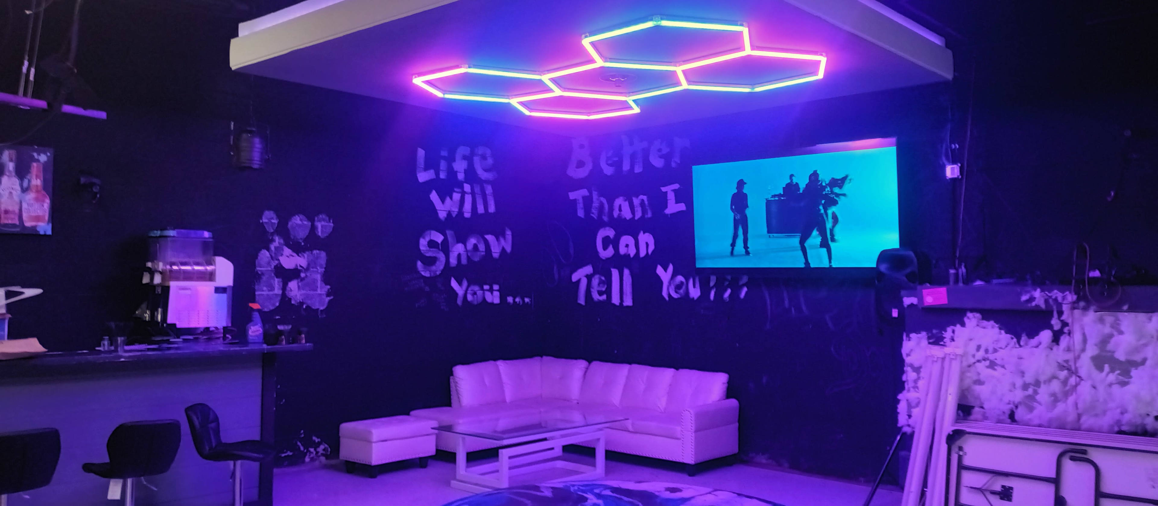 A room features a white couch and a glowing hexagonal light fixture, with neon walls displaying inspirational phrases and a large television screen showing a video.