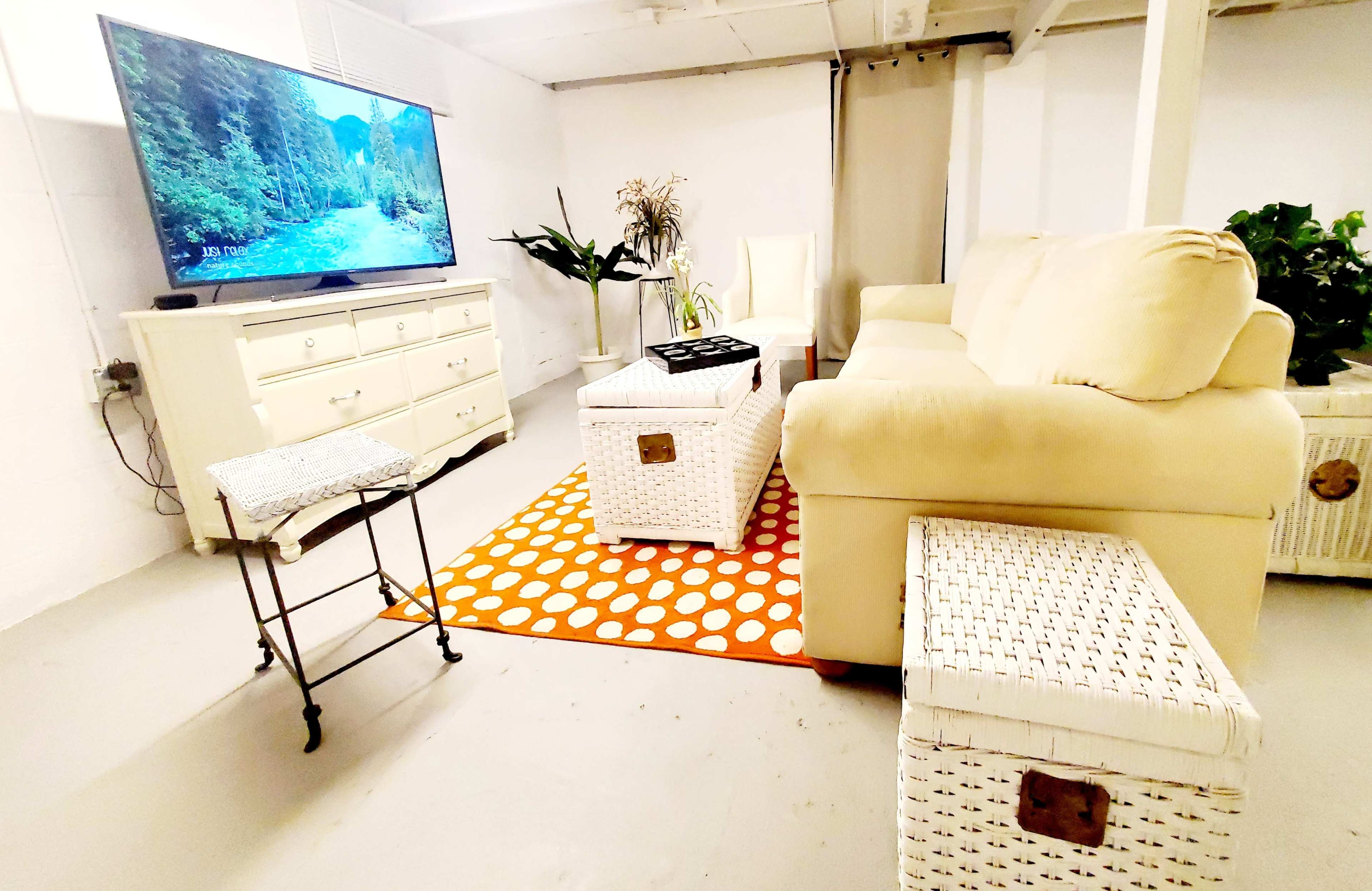 A furnished basement living area features a light-colored couch, a television mounted on the wall, and a decorative area rug with a pattern of orange and white polka dots.