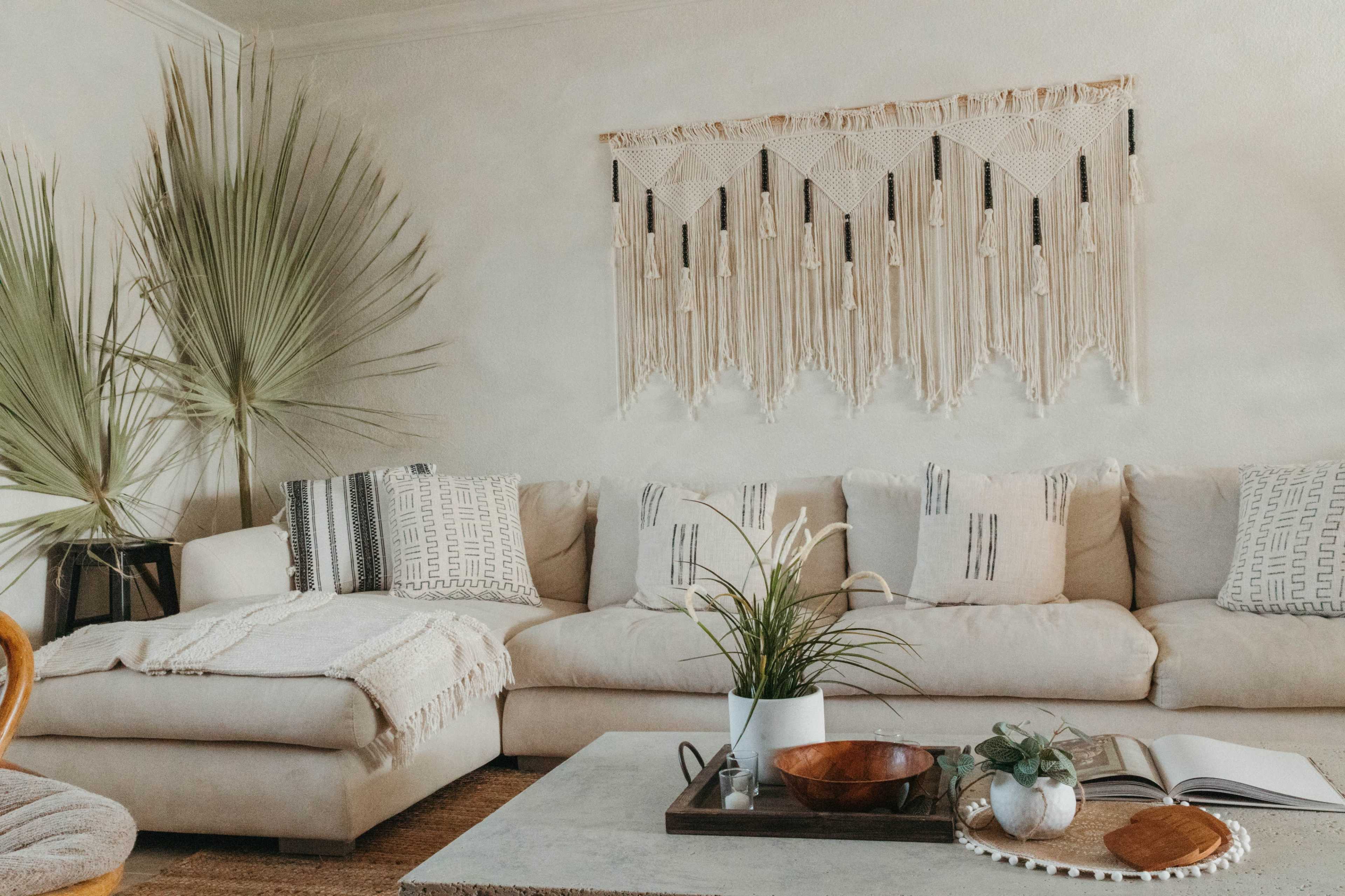 A cozy living room with a beige sofa adorned with patterned pillows, a macramé wall hanging, and decorative plants beside a coffee table.