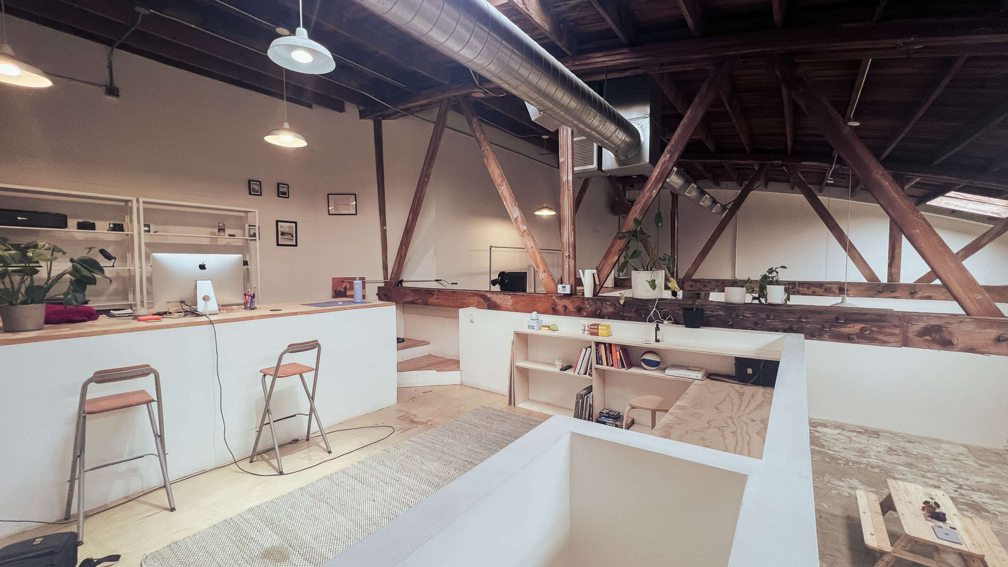The image shows a spacious, modern office interior with wooden beams, a minimalist design, and a layout that includes a kitchen area and workstations.