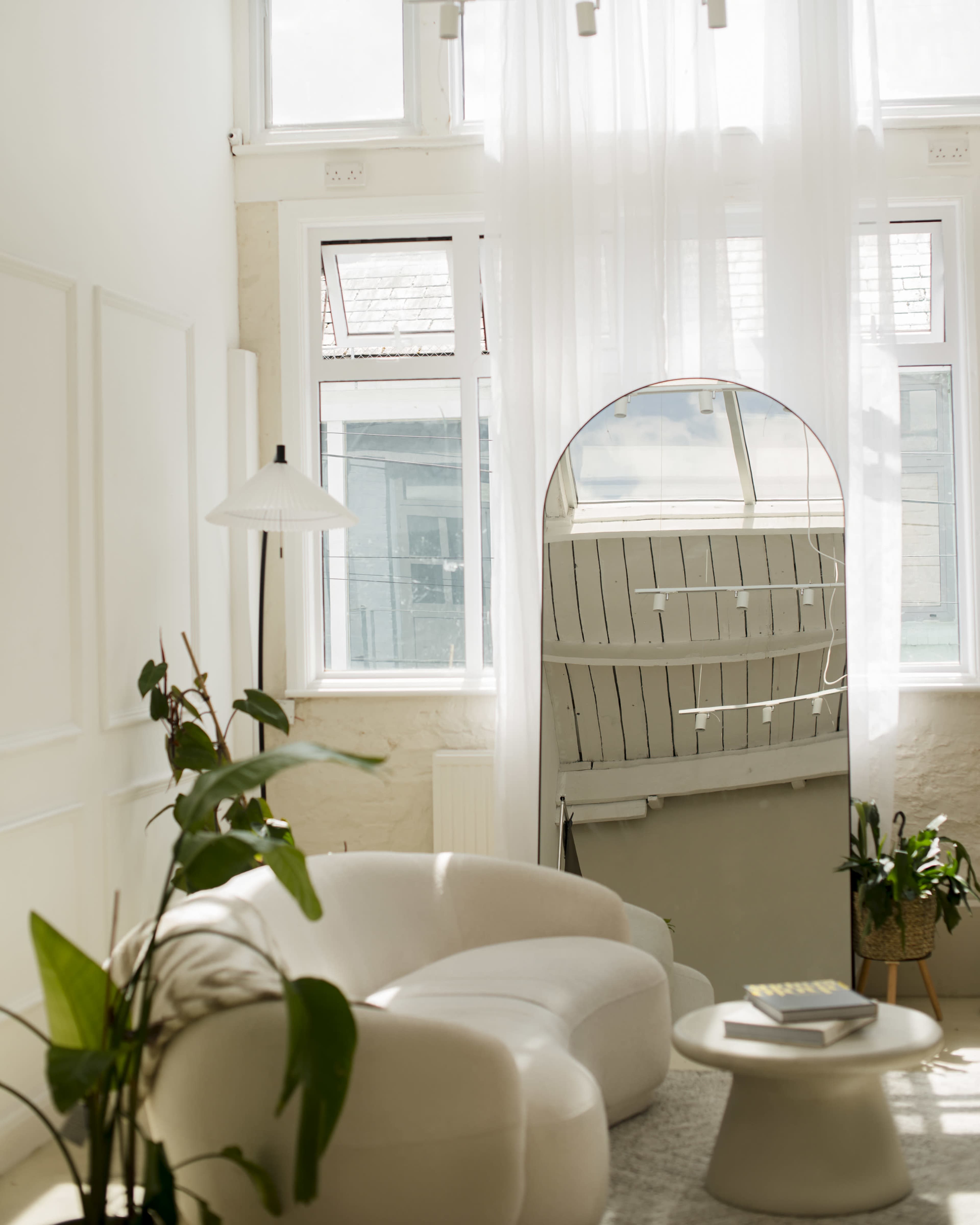 The image shows a brightly lit, modern living room featuring a white curved sofa, a round coffee table, a floor lamp, and large windows with sheer curtains.