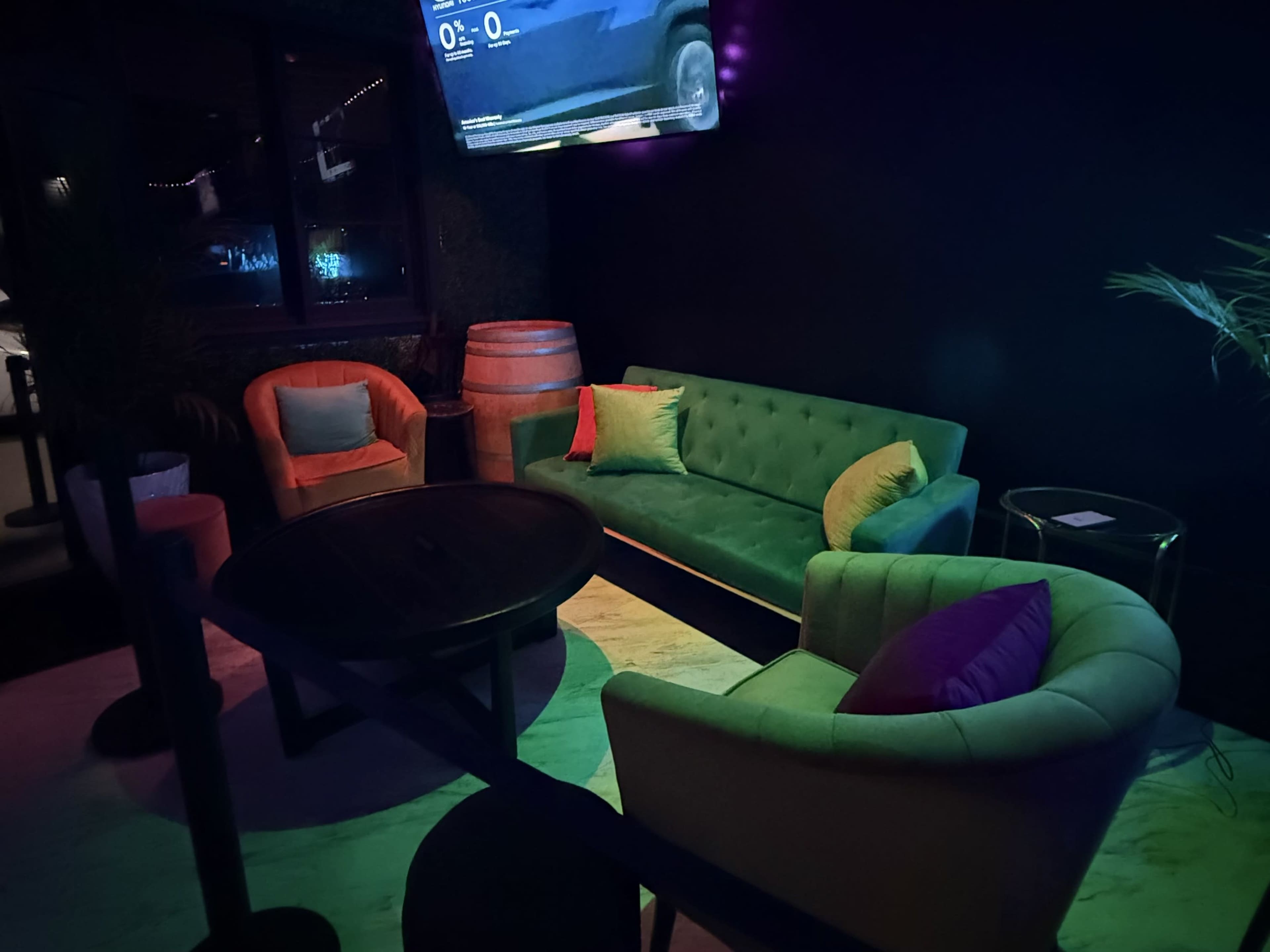 The scene features a cozy seating area with a green sofa and two armchairs, set against a dark wall with a flat-screen TV above and a barrel in the background.