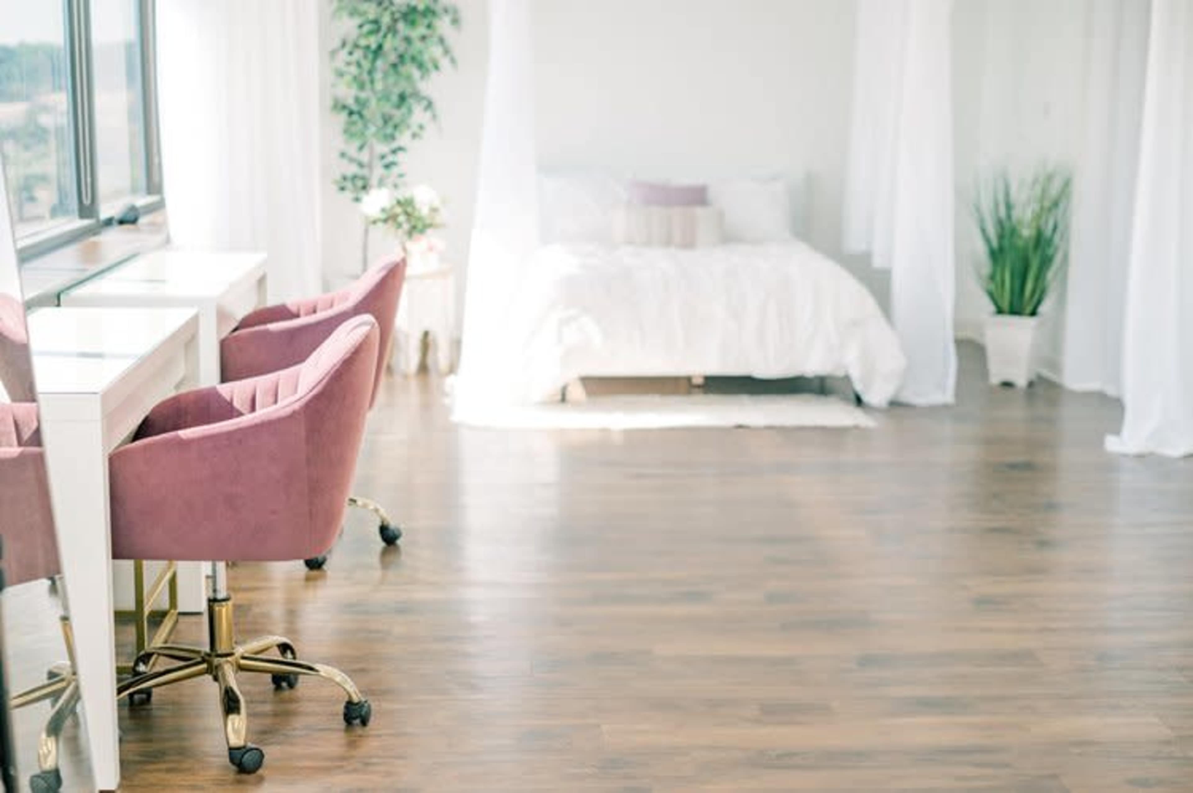 The image shows a bright, modern bedroom with a white bed, pink chairs at a desk, and potted plants.