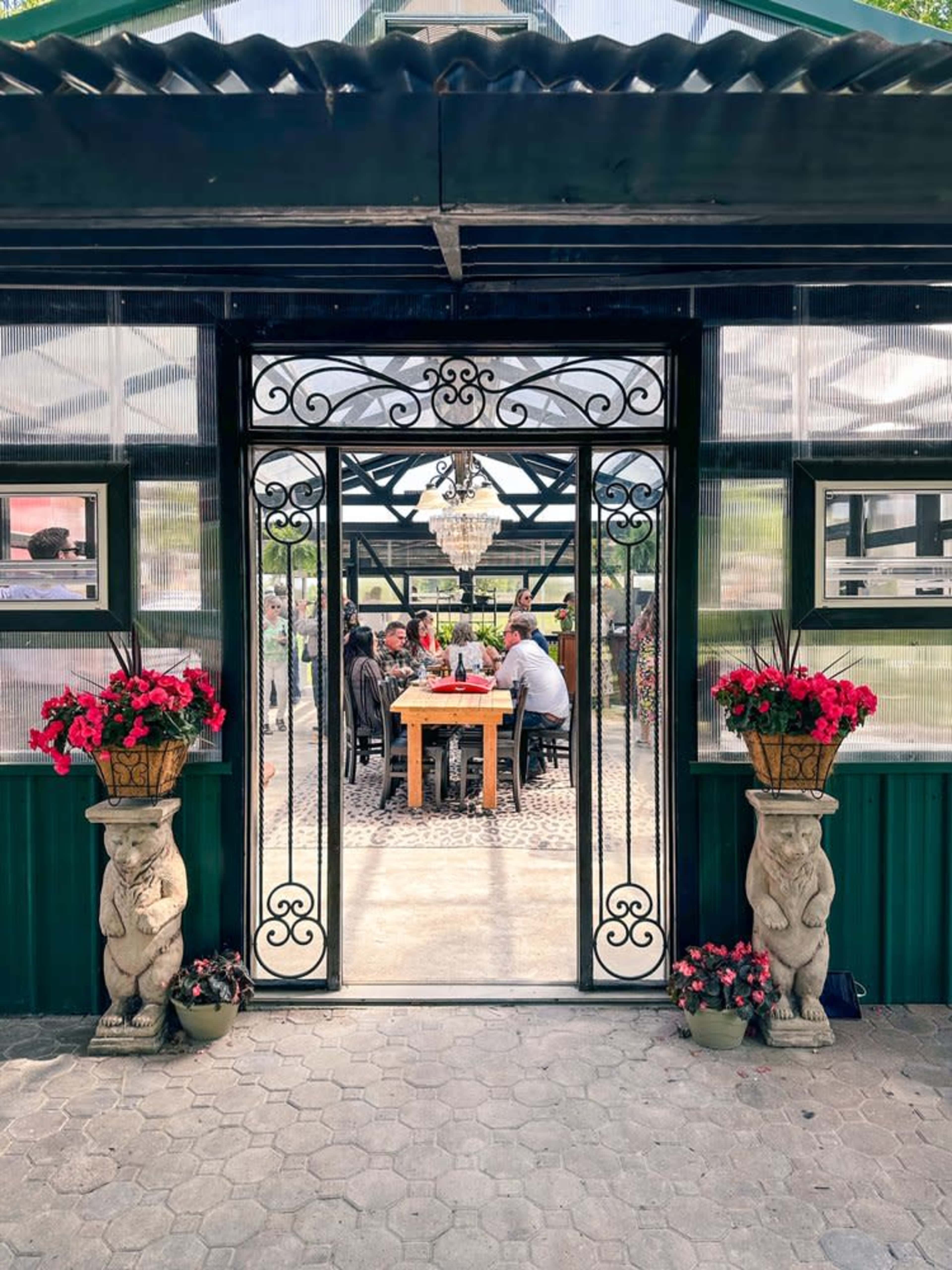 A glass-walled greenhouse features a dining area with a chandelier, flanked by potted flowers and decorative stone sculptures at the entrance.