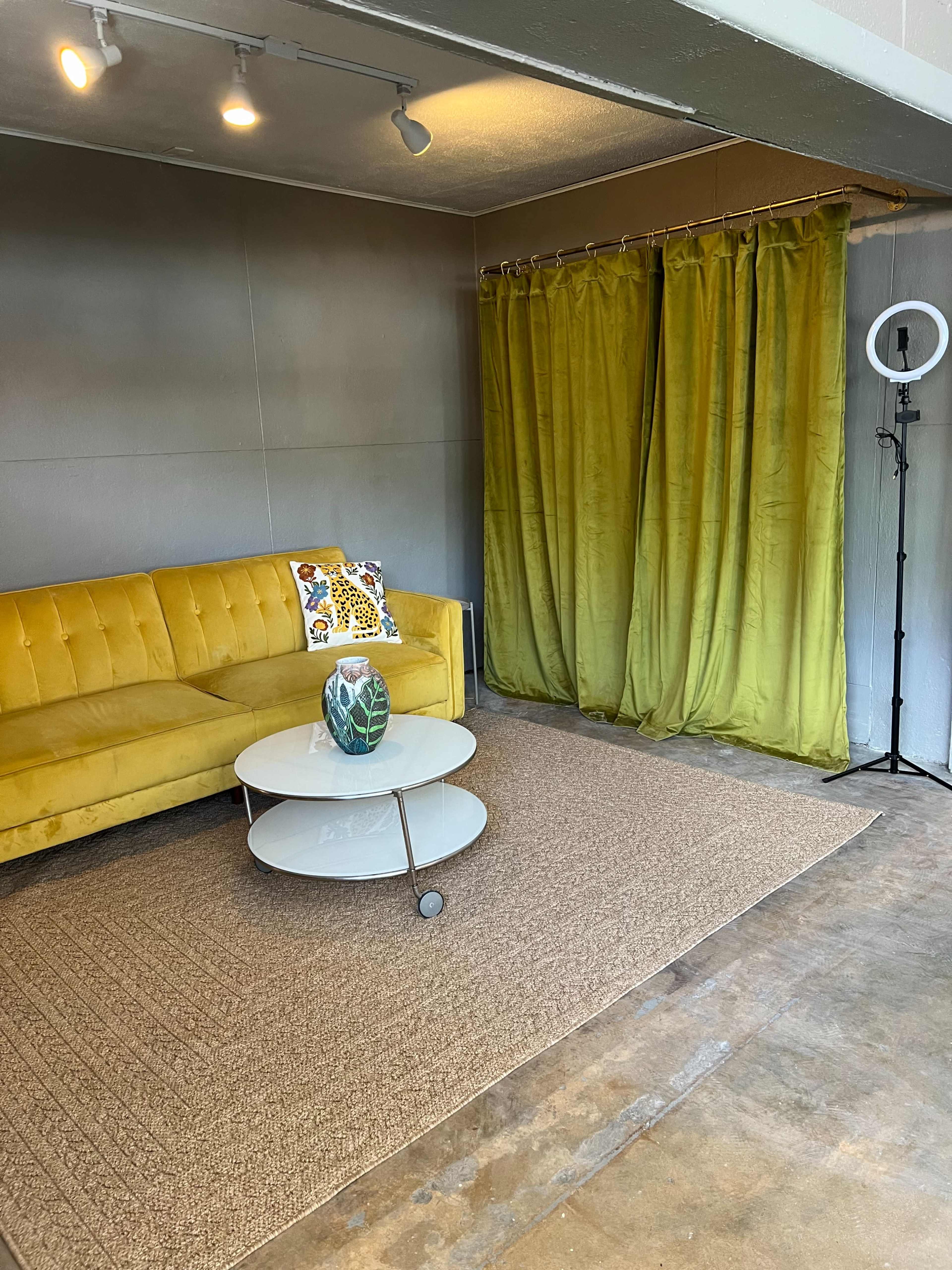 The image shows a brightly lit room featuring a yellow couch, a circular glass coffee table on a textured rug, and a green curtain along with a ring light in the corner.