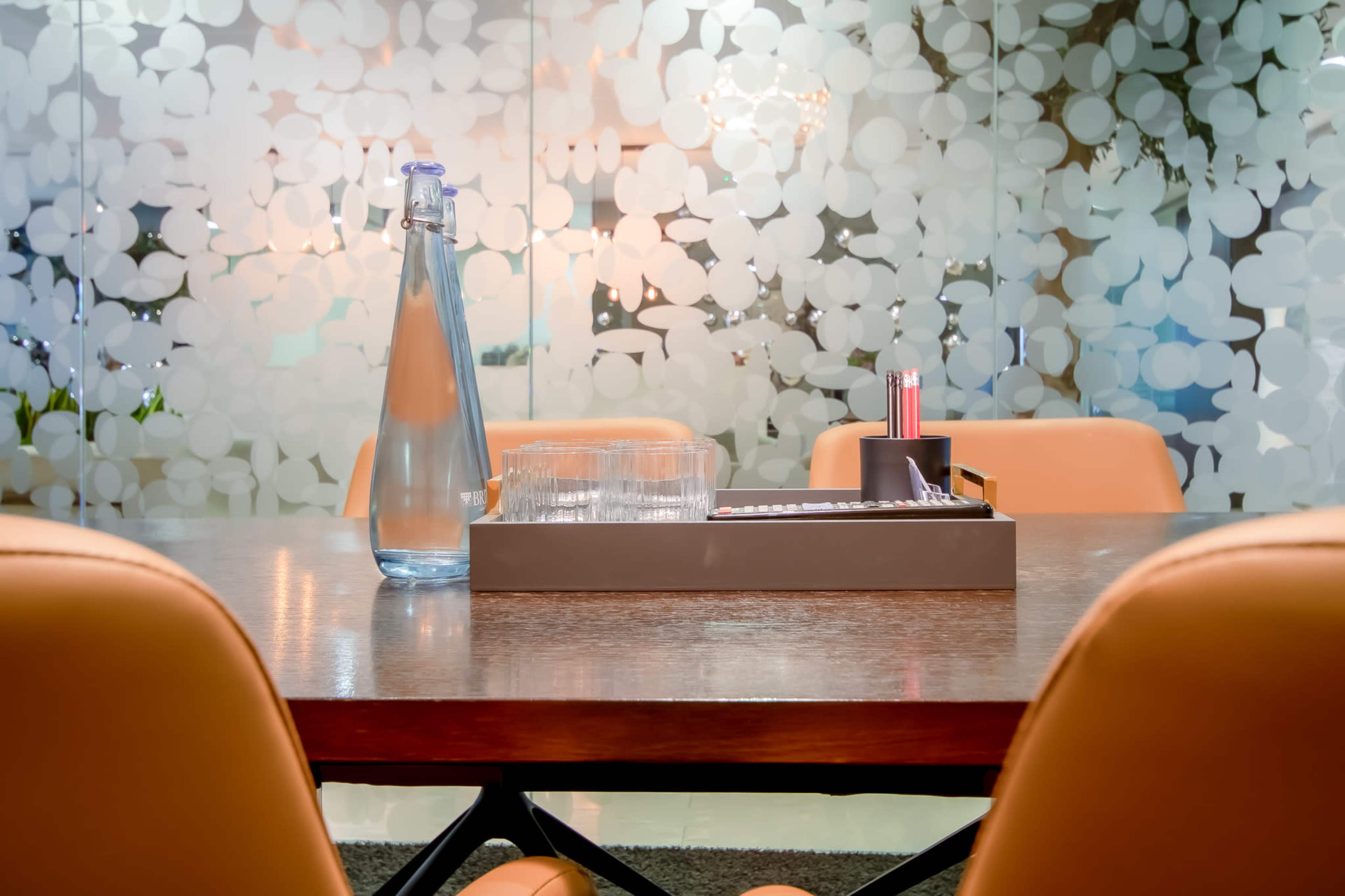 A water carafe and glasses are placed on a tray at a wooden table surrounded by orange chairs in a room with frosted glass walls.