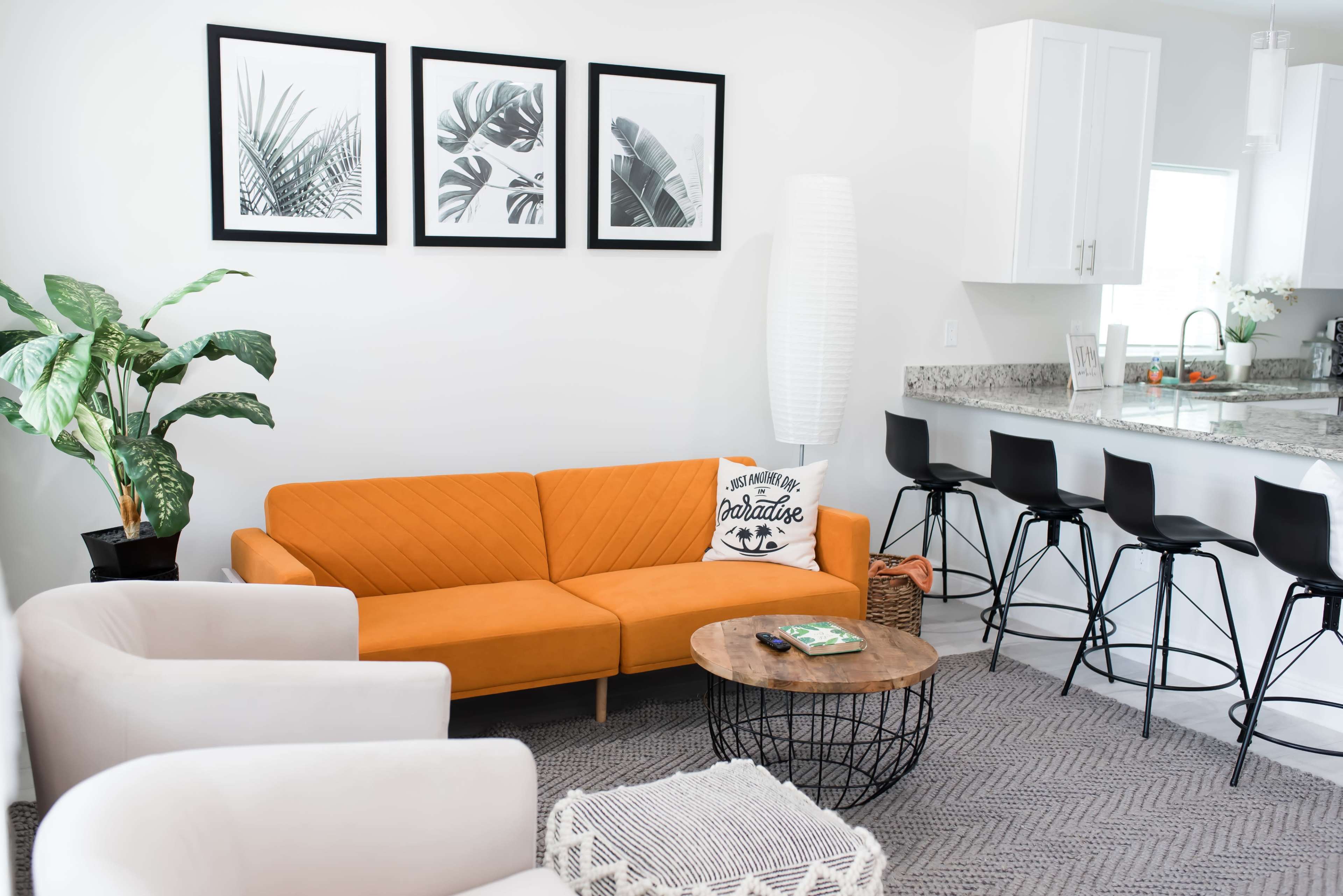 A modern living room features an orange couch, two white armchairs, a round coffee table, and a kitchen bar with black stools, all set against a backdrop of framed botanical prints.