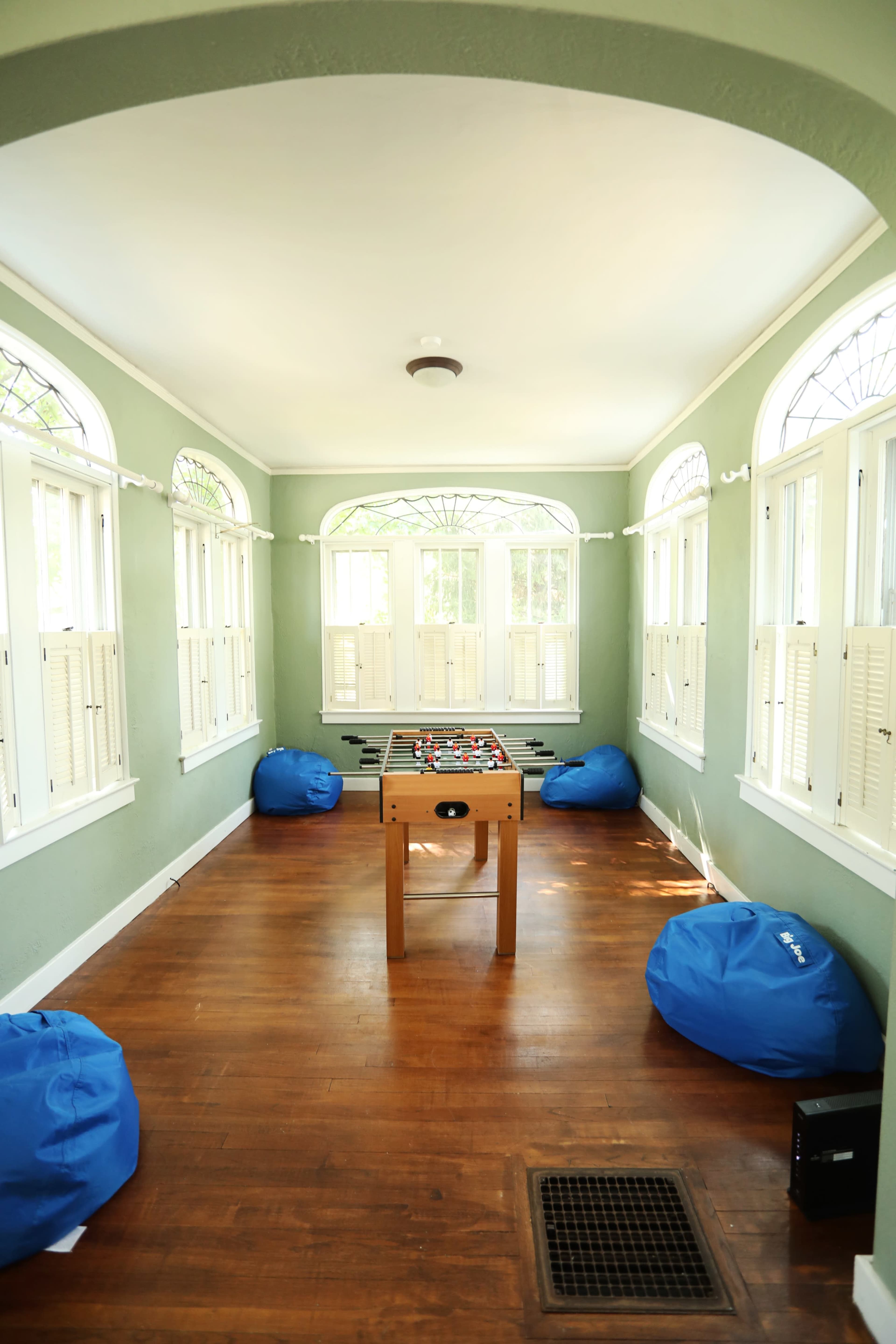 The image shows a bright room with large windows, a foosball table in the center, and three blue bean bag chairs arranged around it on a wooden floor.