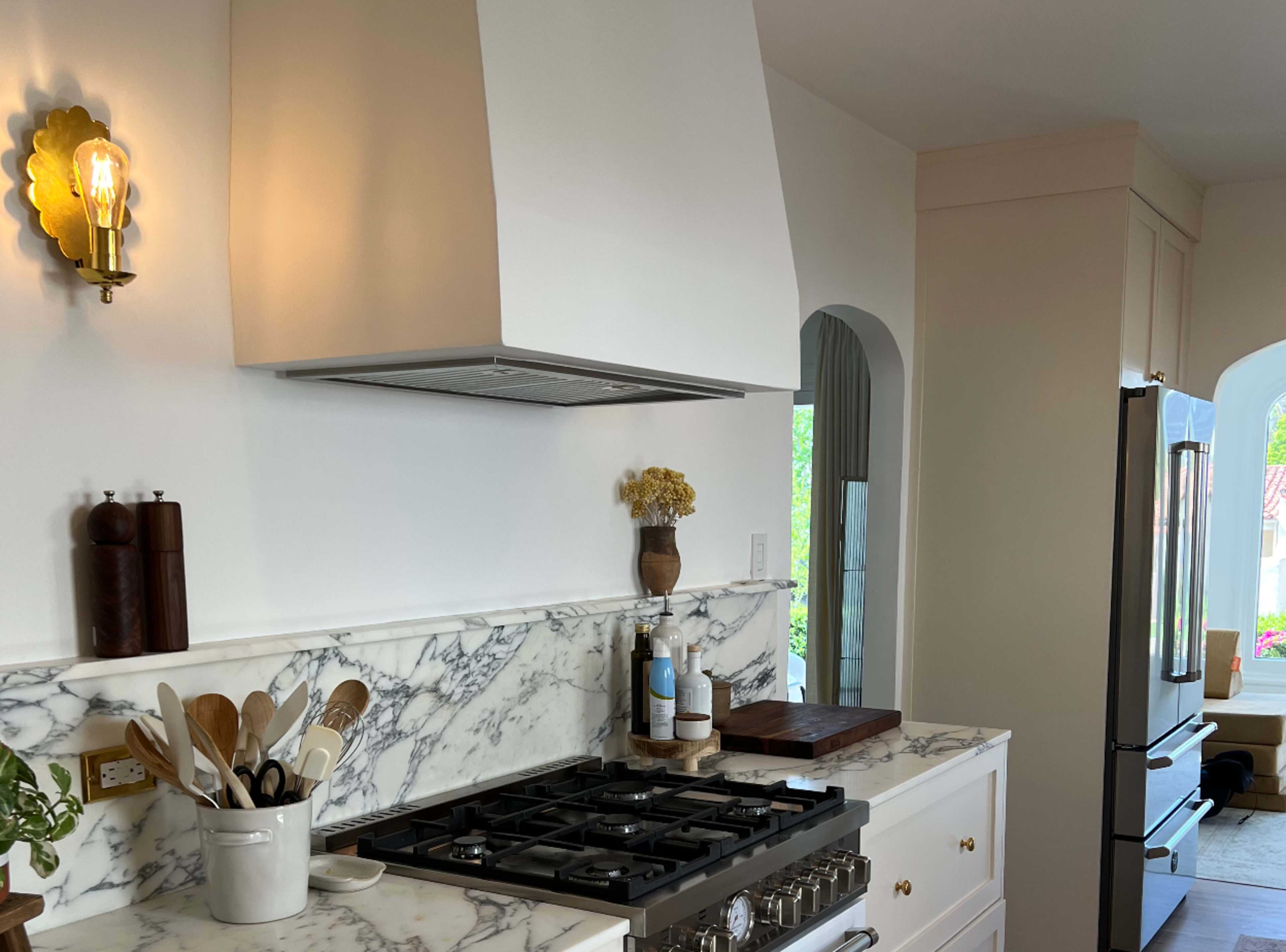 The image shows a modern kitchen with a marble countertop, a stainless steel range hood, and various cooking utensils displayed near a gas stove.