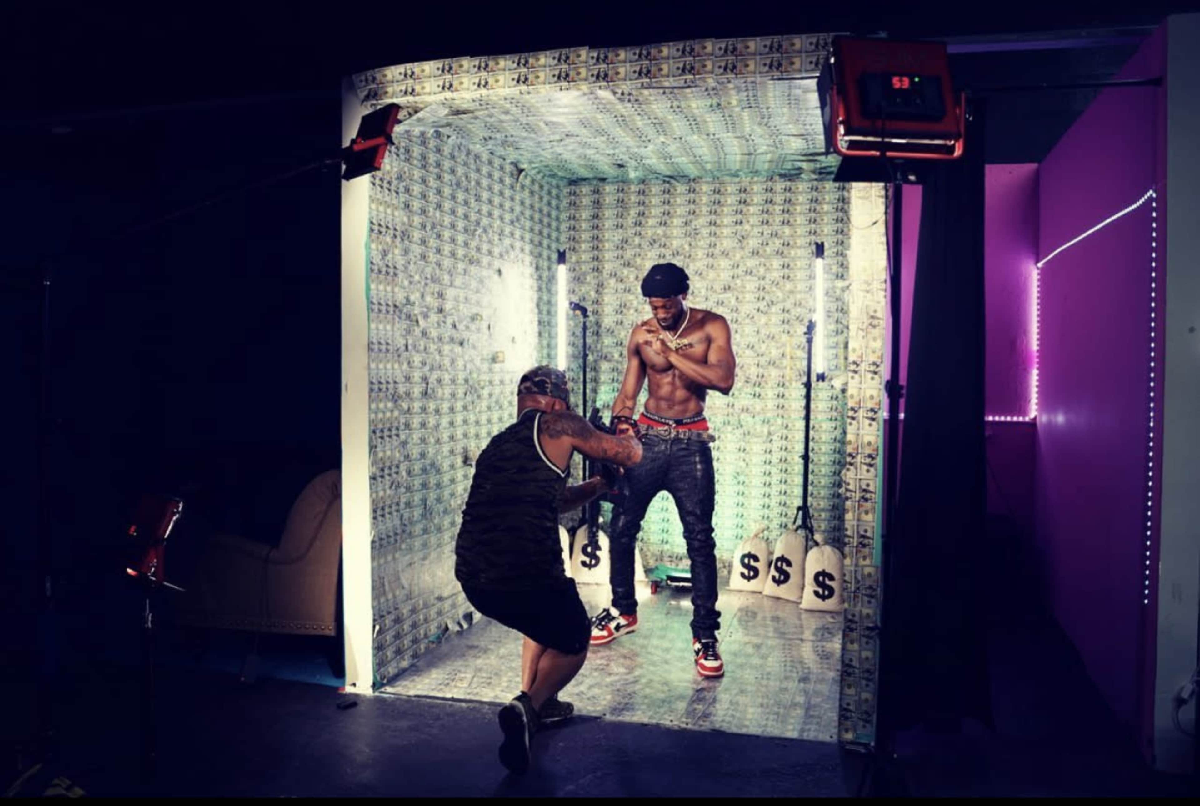 A shirtless man poses inside a room decorated with money-themed walls while another person adjusts the lighting.