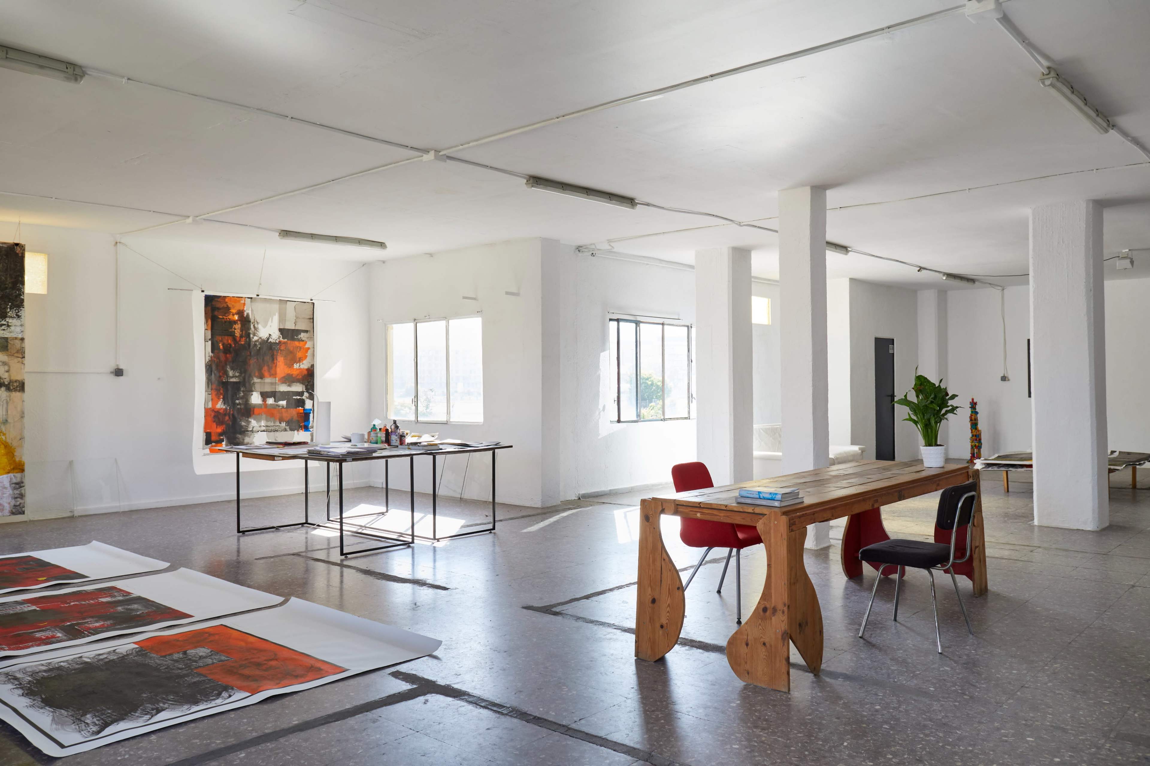 An artist's studio with large windows, a wooden table, red chairs, and artwork displayed on the floor and walls.