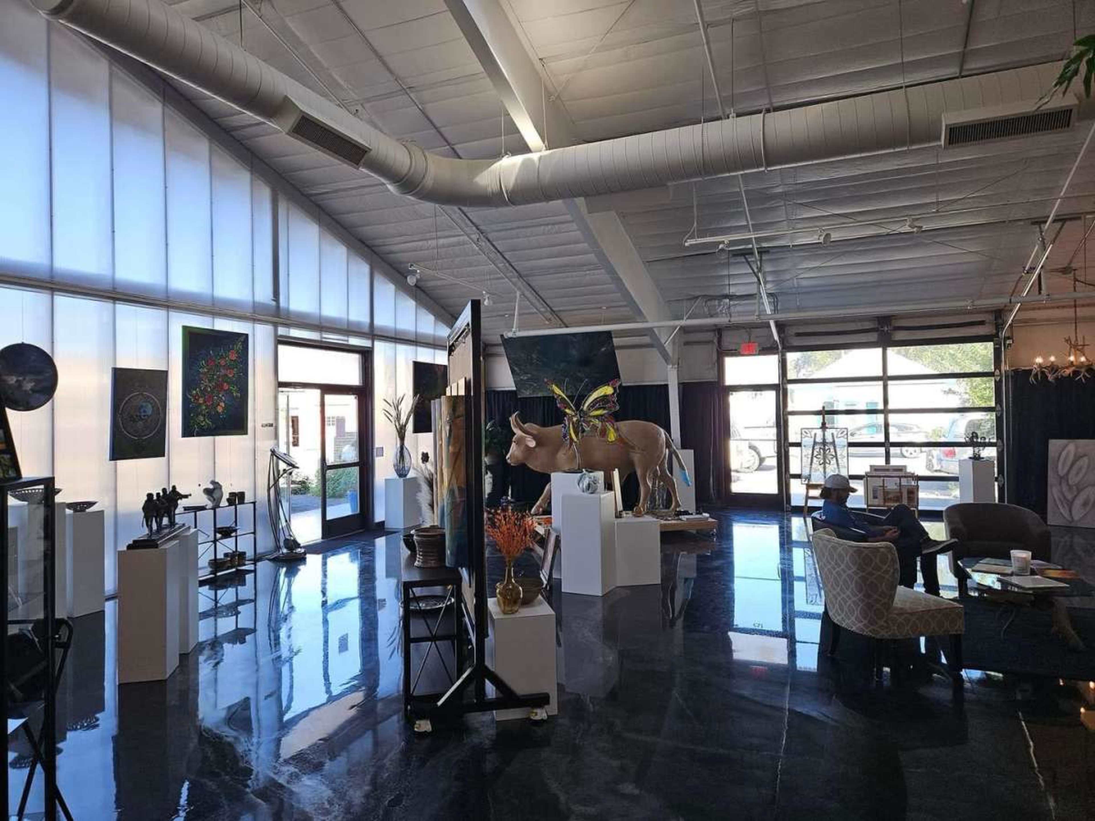The image shows an art gallery featuring various sculptures and paintings displayed on pedestals and walls in a spacious, well-lit interior.