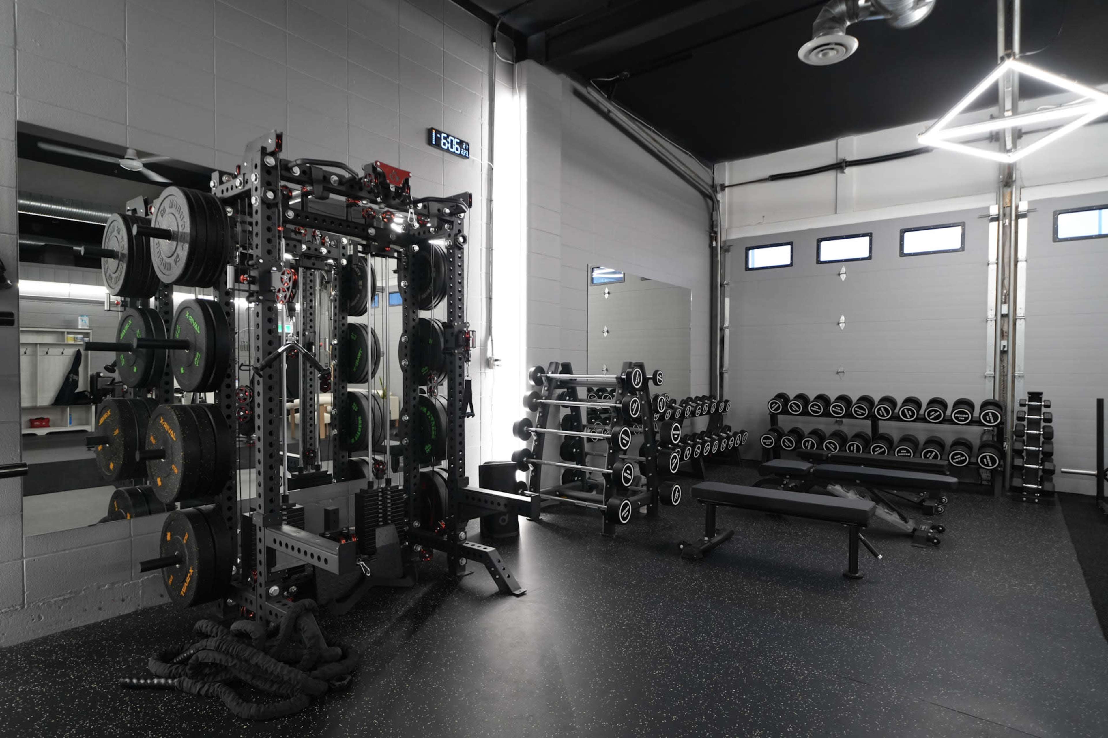 The image shows a modern gym with a weightlifting station, a mirrored wall, and organized free weights on racks.