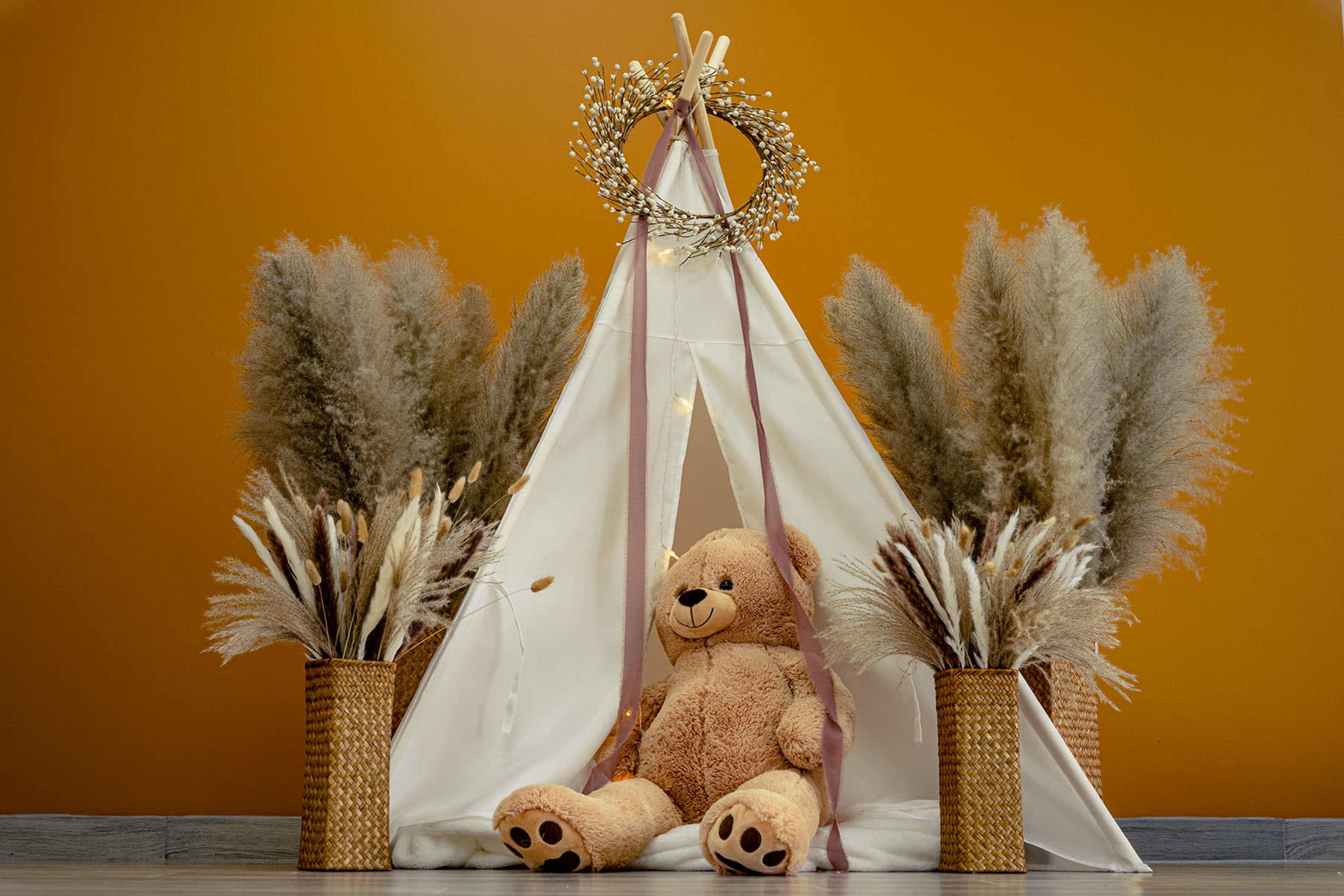 A stuffed teddy bear sits inside a white teepee tent, flanked by two large bouquets of dried grass in woven baskets against an orange wall.