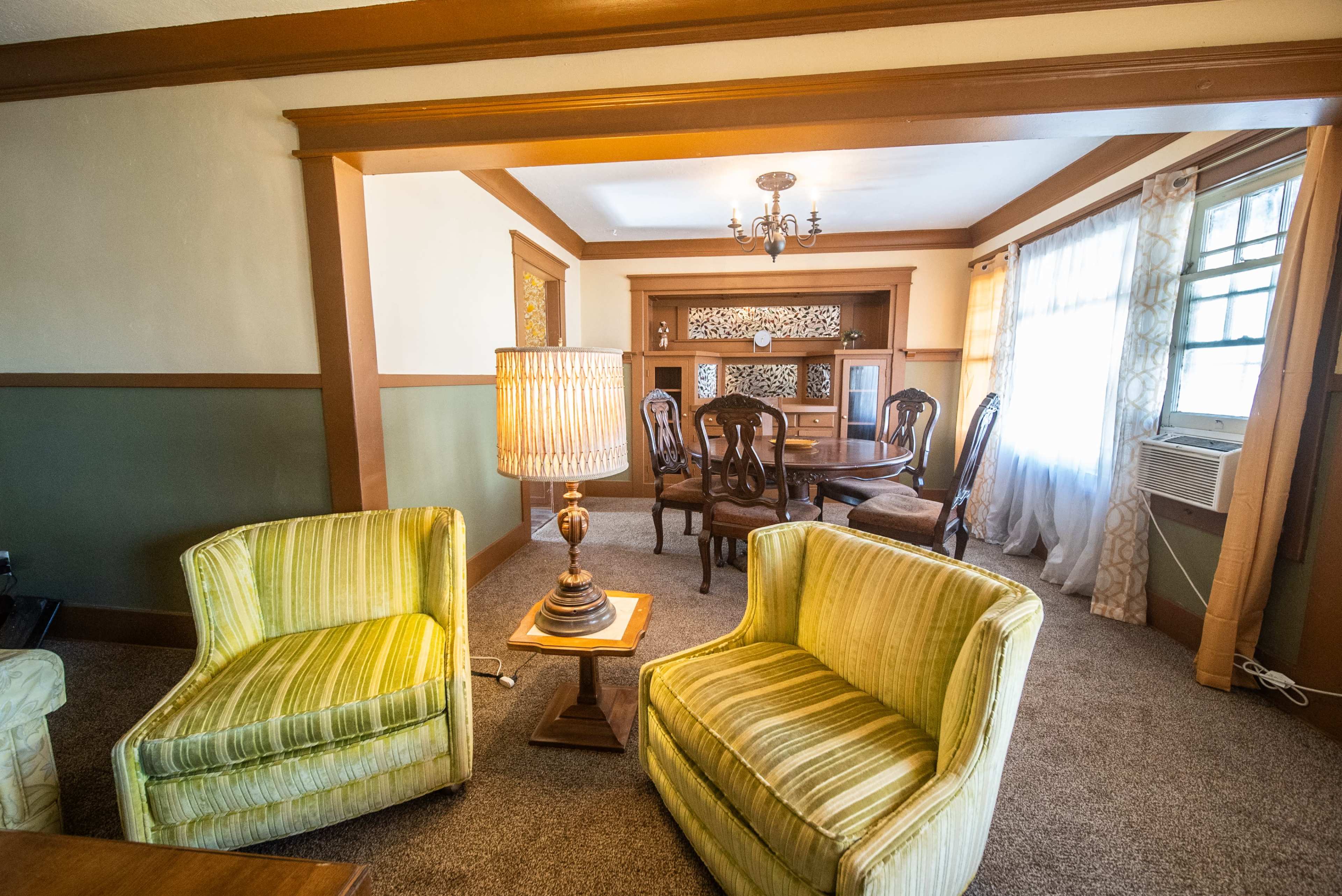 A living room with two striped armchairs, a table lamp, and a view into a dining area featuring a round table and chairs.