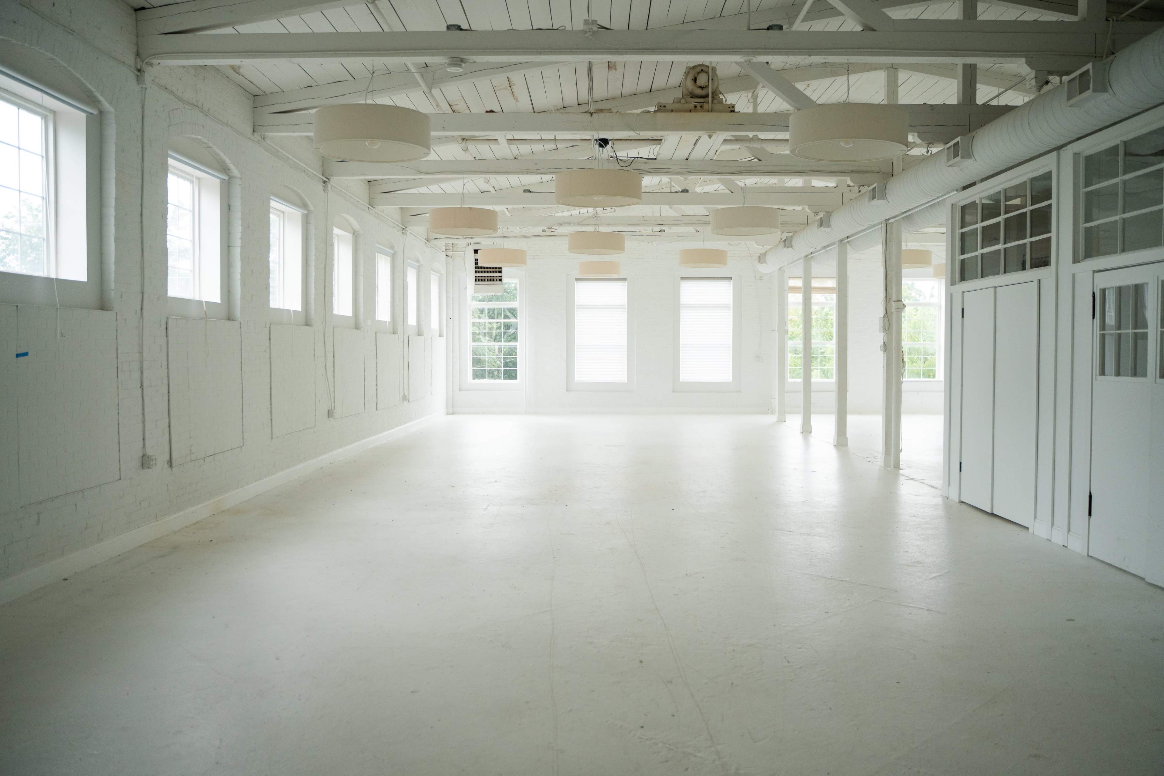 The image shows a spacious, empty room with white walls, large windows, and overhead lighting.