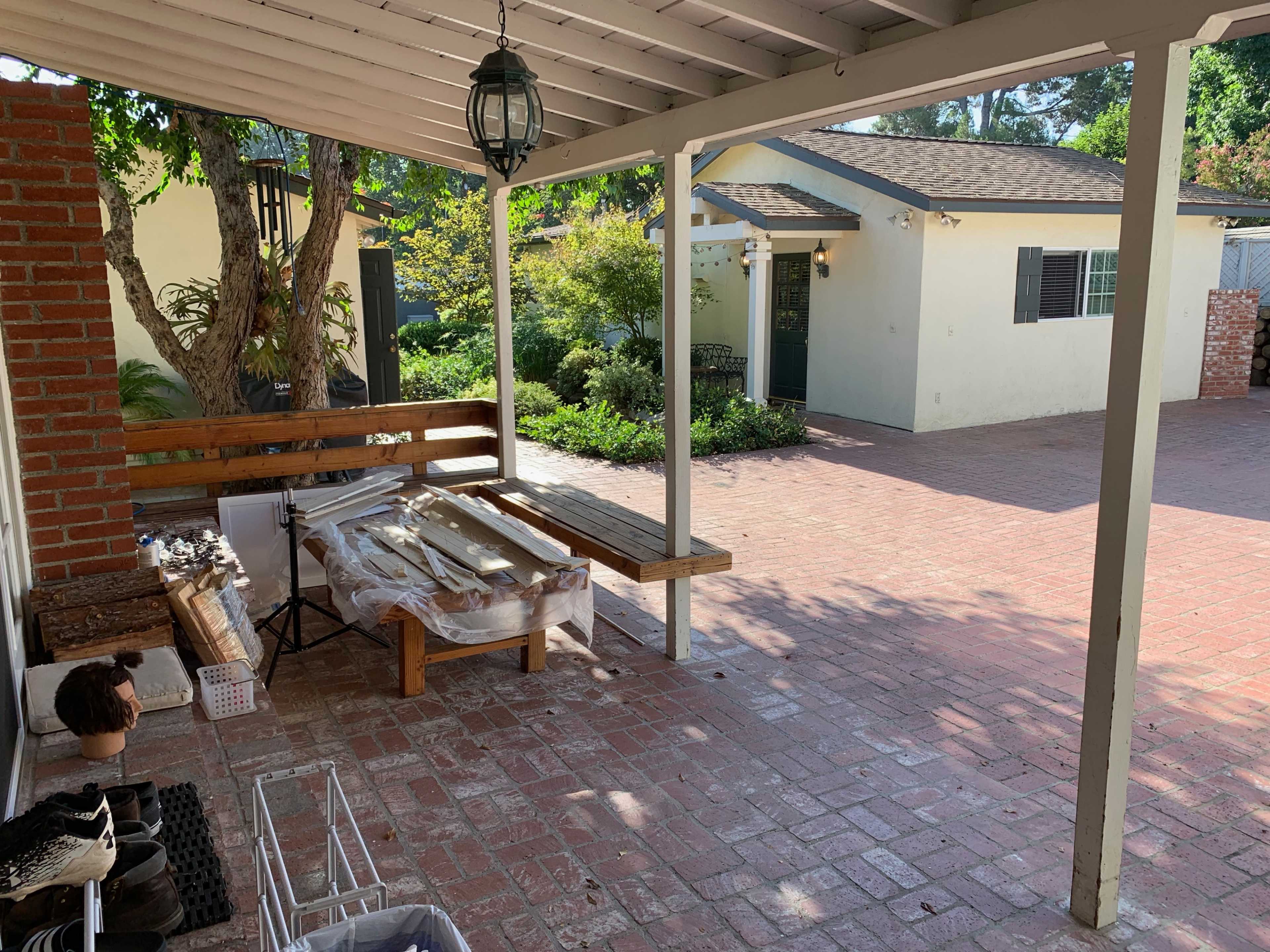 A covered porch area features a wooden bench and various items laid out on a table, with a garden and a house visible in the background.