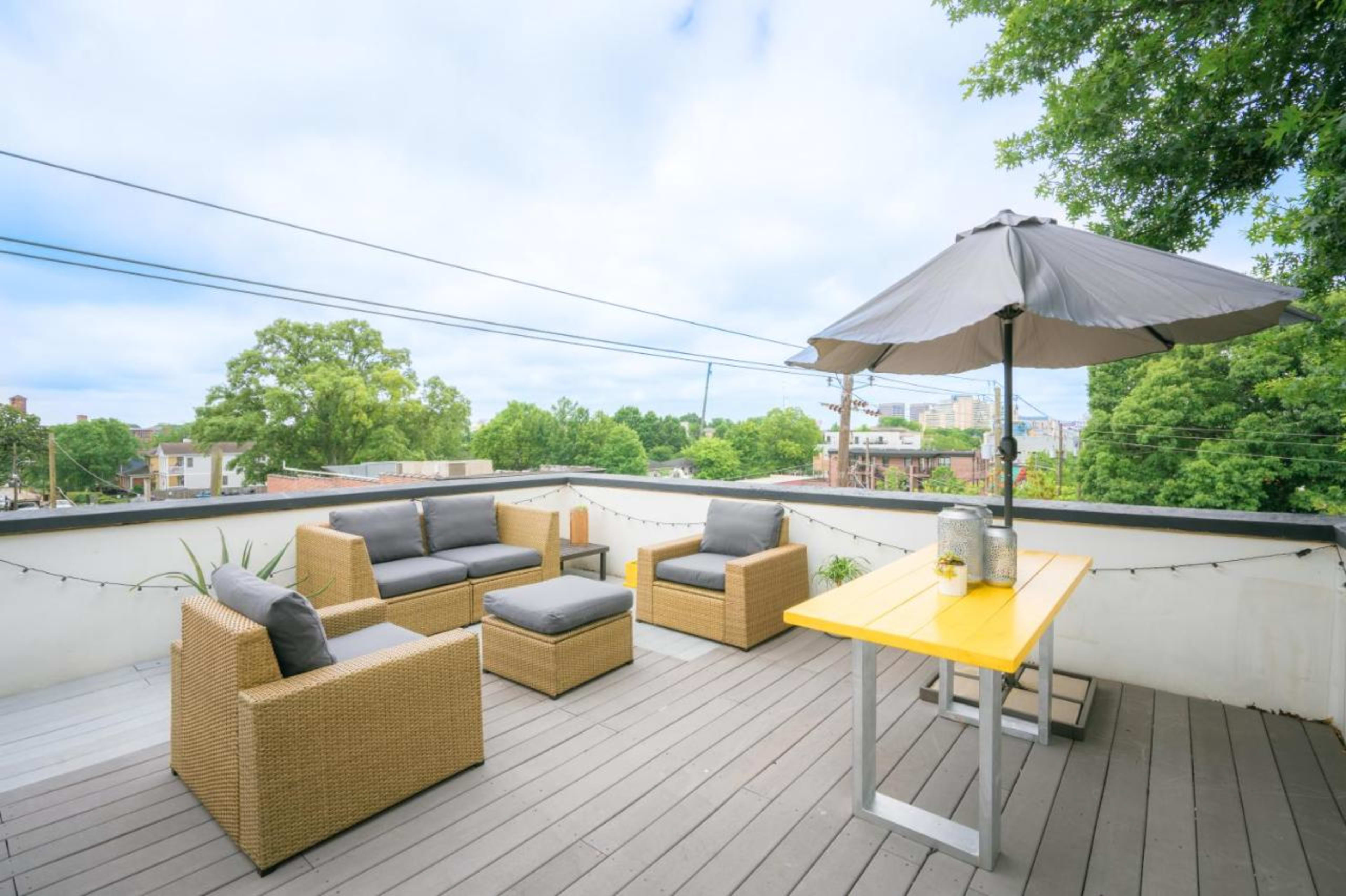 The scene features a rooftop terrace with wicker seating, a yellow table, and an umbrella, surrounded by greenery and city buildings in the background.