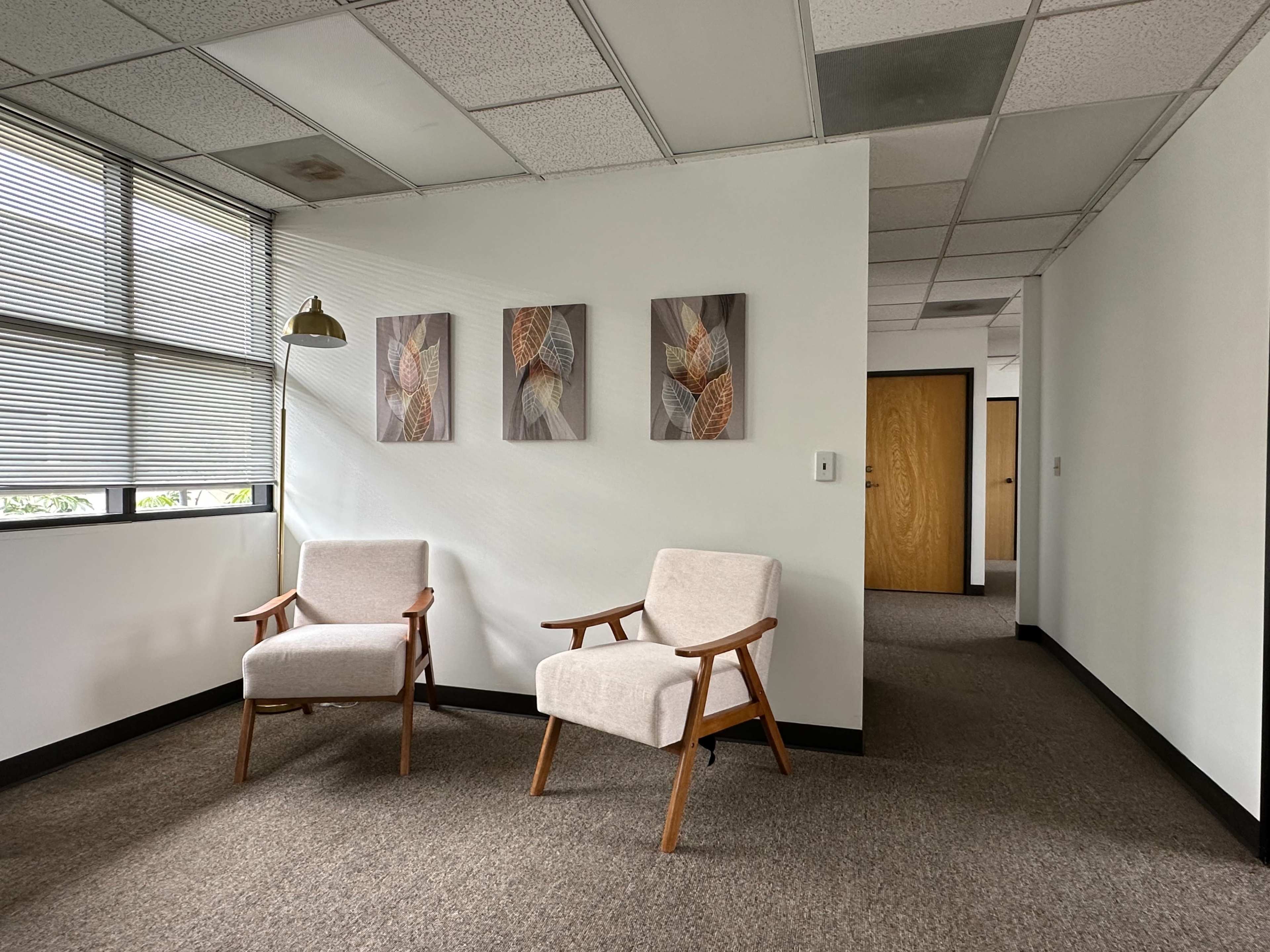 A small office space with two light-colored chairs placed next to a floor lamp, and three framed artworks on the wall.