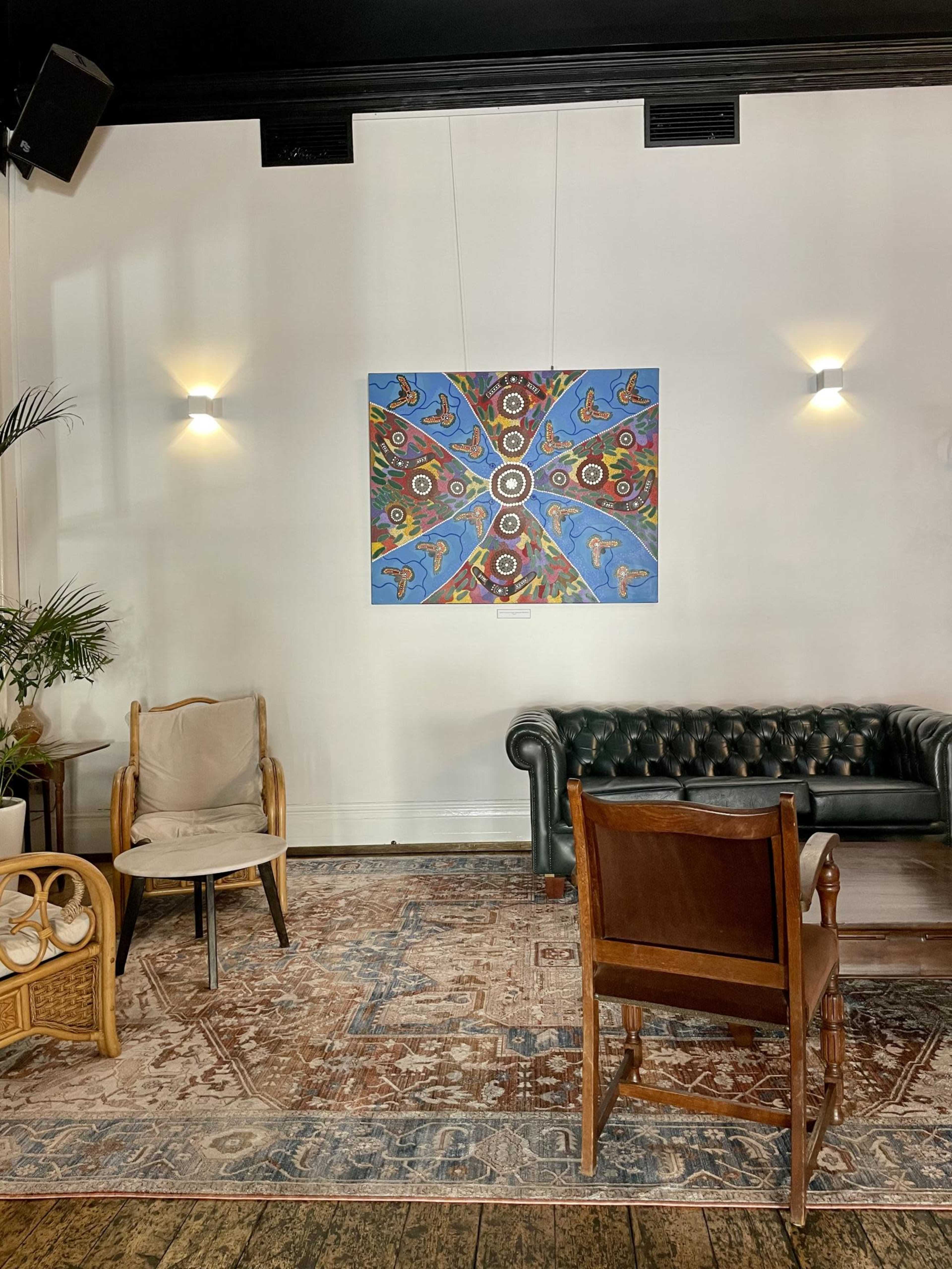 The image shows a cozy lounge area with a colorful abstract painting on the wall, a dark leather couch, and wooden chairs arranged on a patterned rug.