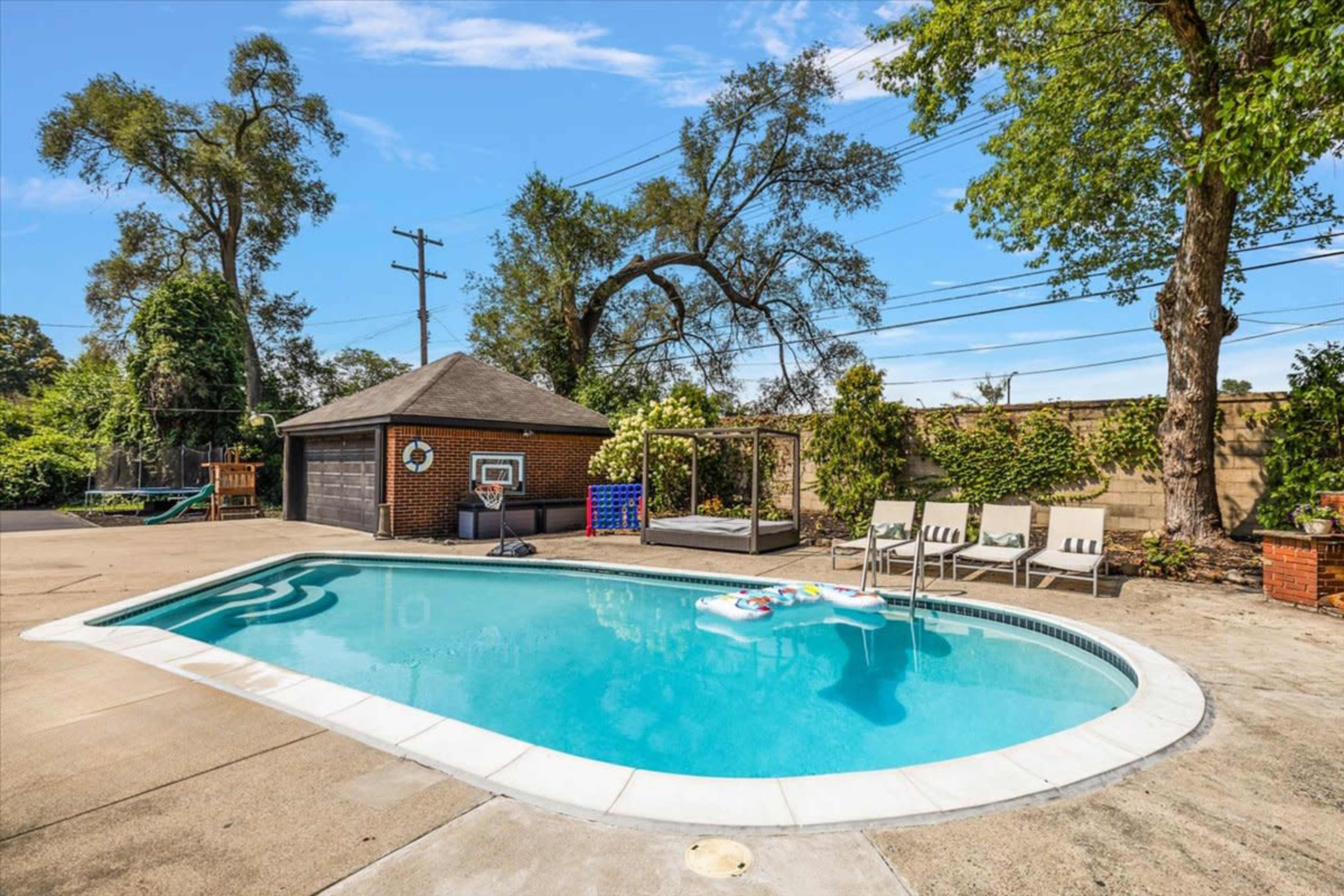 The image shows a backyard swimming pool surrounded by a patio, lounge chairs, and a storage shed, with tall trees and a fence in the background.