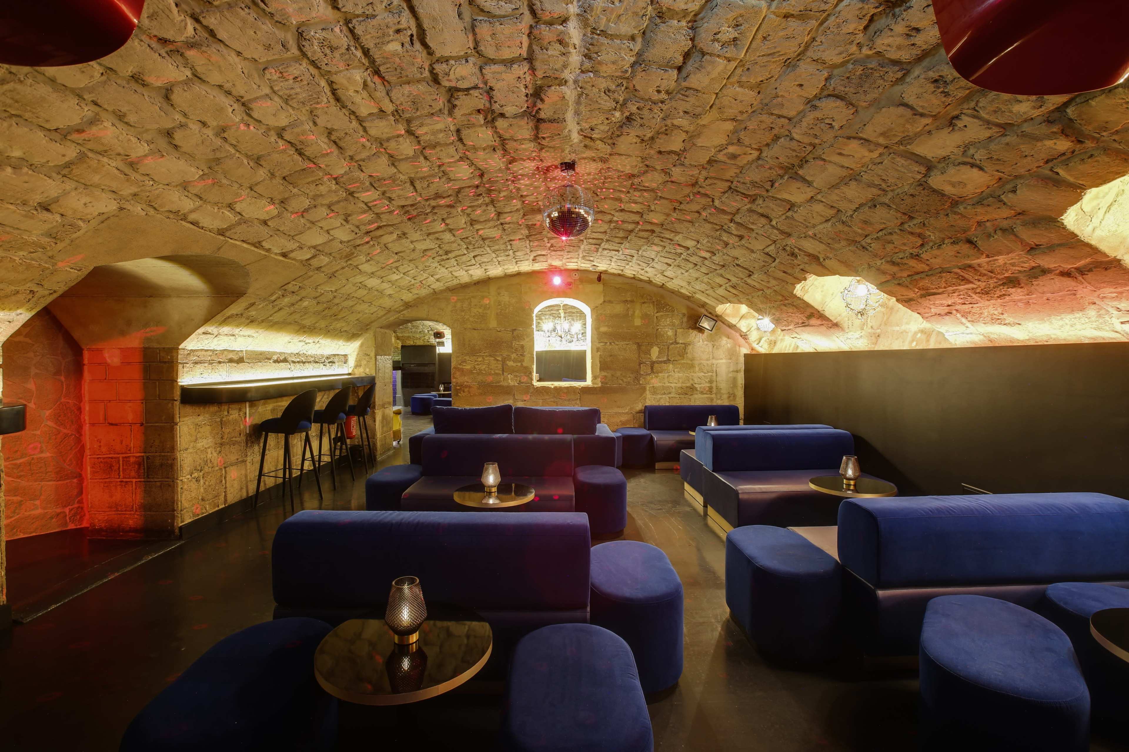A dimly lit lounge with arched stone ceilings, blue seating arrangements, and a bar area in the background.