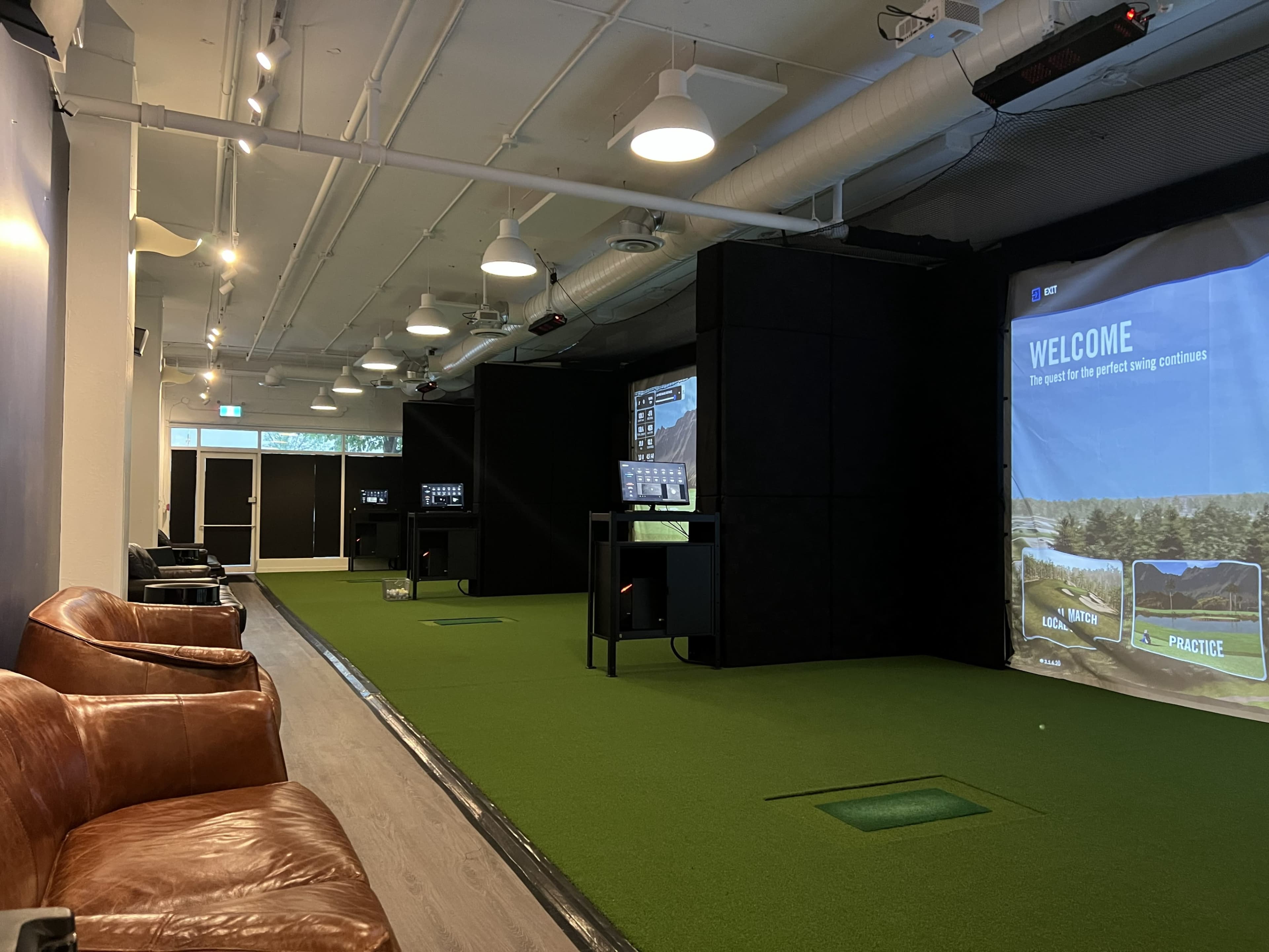 The image shows an indoor golf simulator facility with green turf, multiple screens displaying golf-related graphics, and brown leather seating.