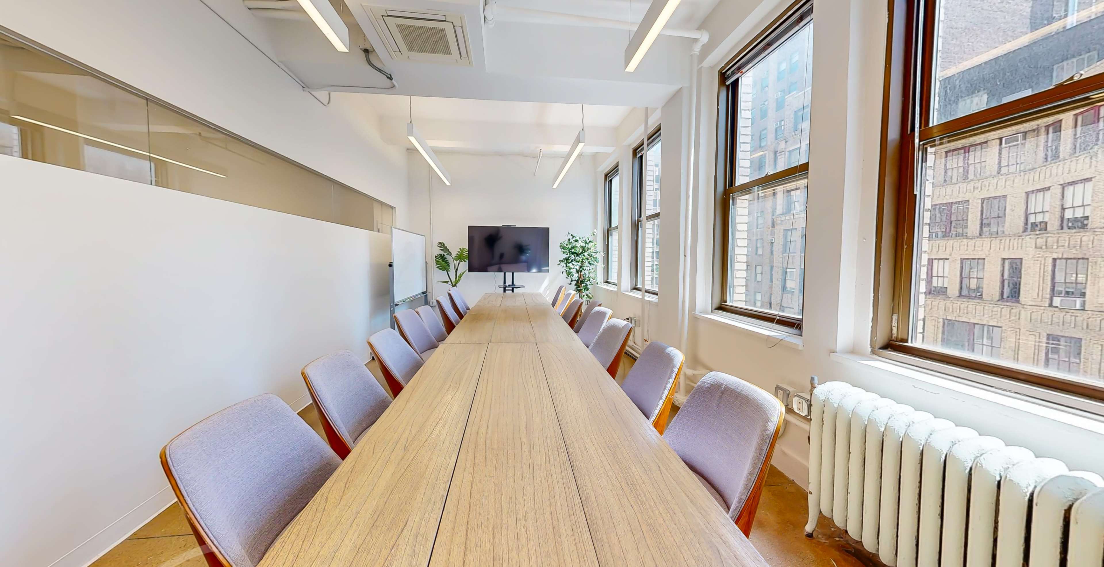 A long wooden conference table is surrounded by chairs in a well-lit meeting room with large windows overlooking a cityscape.