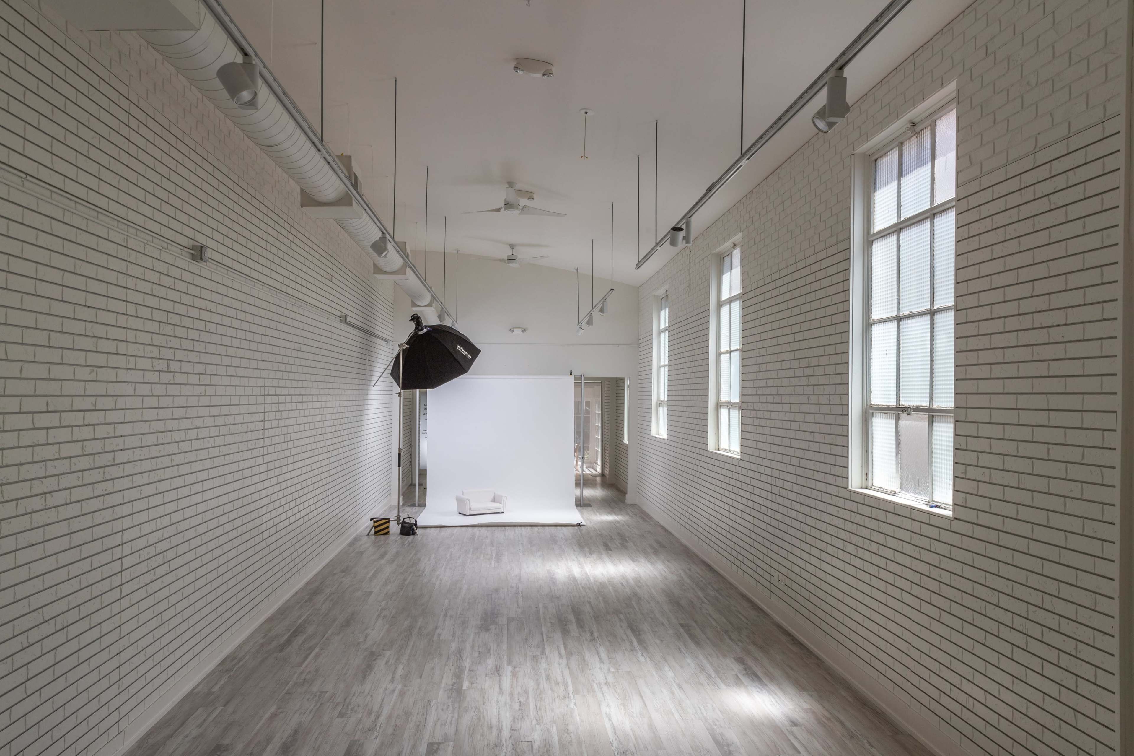 A long, narrow studio space with white brick walls, large windows, and a backdrop setup at one end.